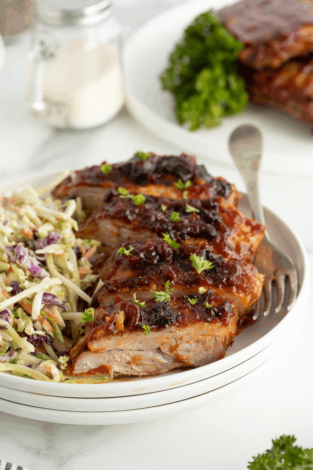 A plate of barbecued pork ribs topped with sauce and herbs is served next to creamy coleslaw. A fork rests on the side, and another plate with ribs and parsley is blurred in the background.