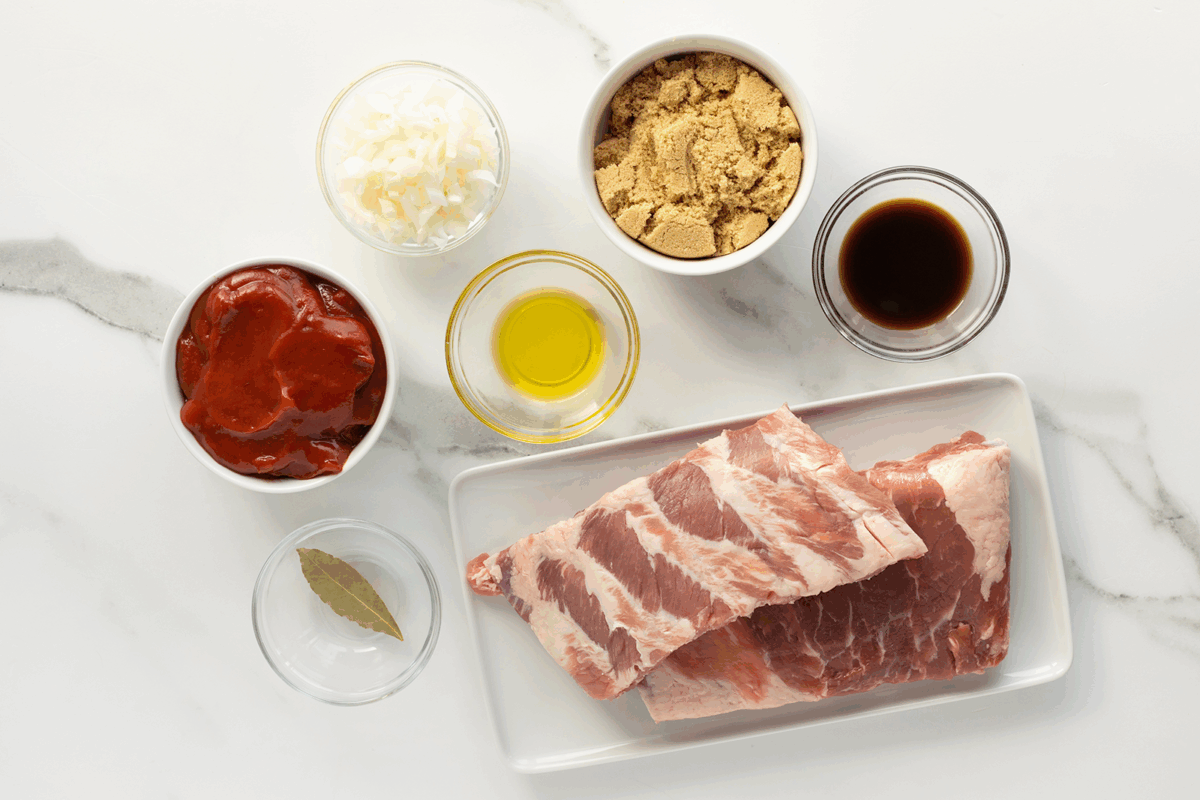 Raw pork ribs on a white plate, surrounded by small bowls containing chopped onions, brown sugar, tomato sauce, olive oil, soy sauce, and a bay leaf, all on a white marble surface.