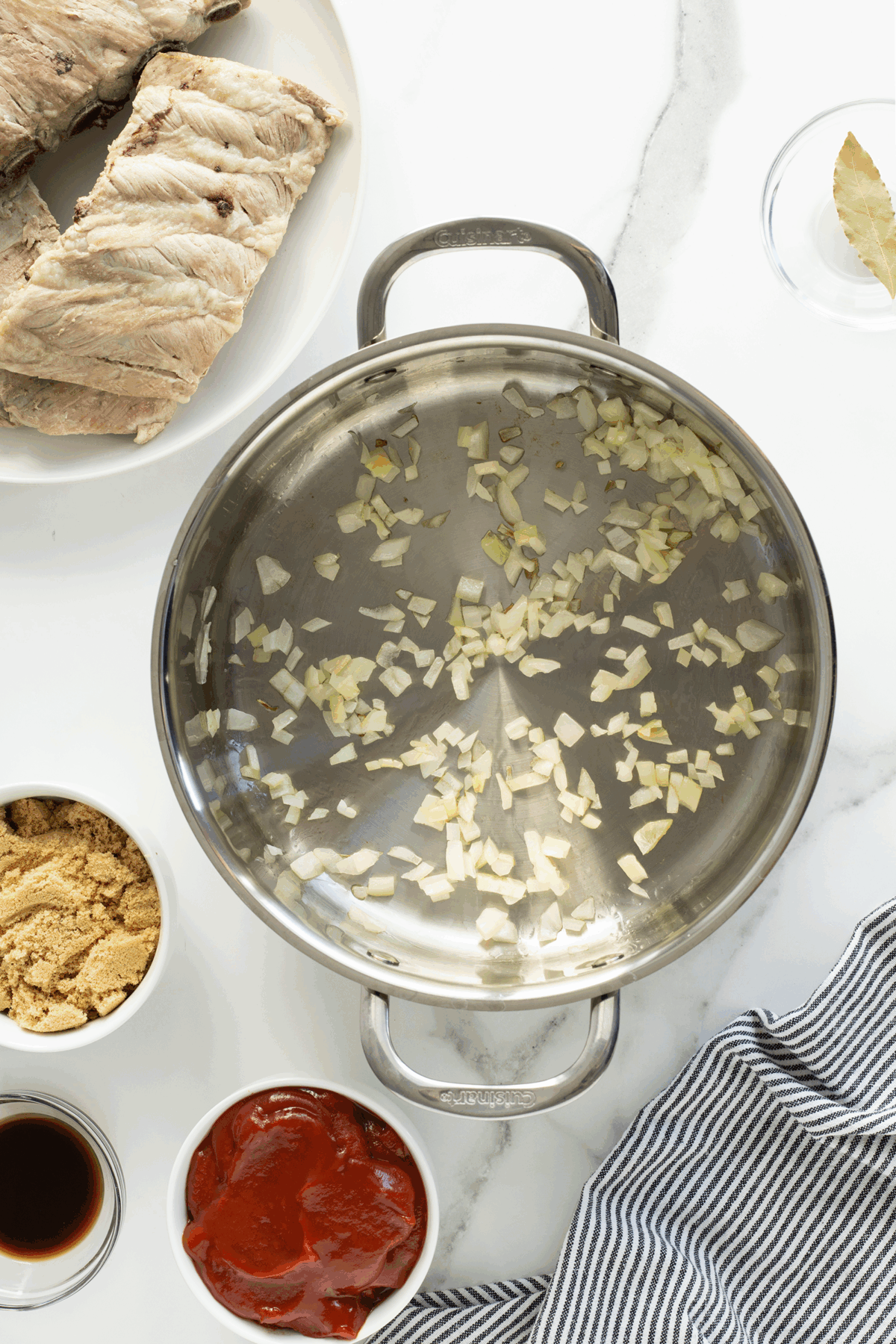 Overhead view of a pot with chopped onions being sautéed. Surrounding the pot are a plate with cooked meat, a bowl of brown sugar, a bowl of tomato sauce, a bowl of soy sauce, a glass with a bay leaf, and a striped cloth.