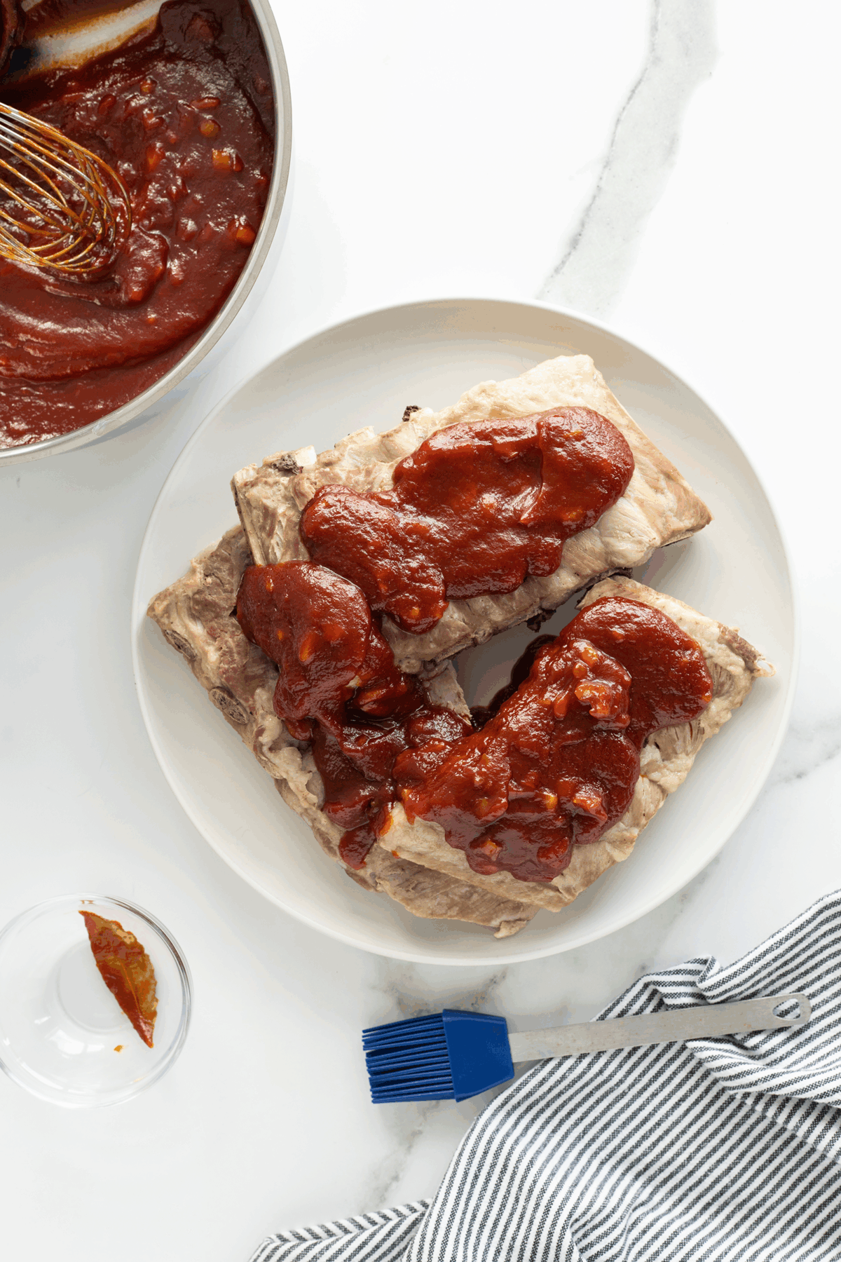 Three pieces of cooked ribs topped with barbecue sauce on a white plate, with more sauce in a pot nearby. A basting brush, empty glass, and striped napkin are also on the white surface.