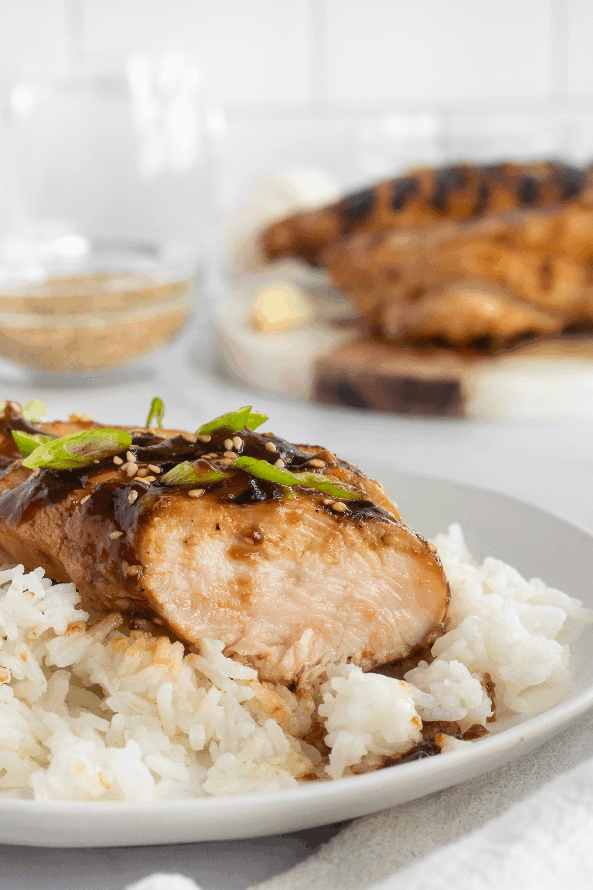A plate of white rice topped with a grilled chicken breast, drizzled with a dark sauce and garnished with sliced green onions. A cutting board with more grilled chicken is blurred in the background.