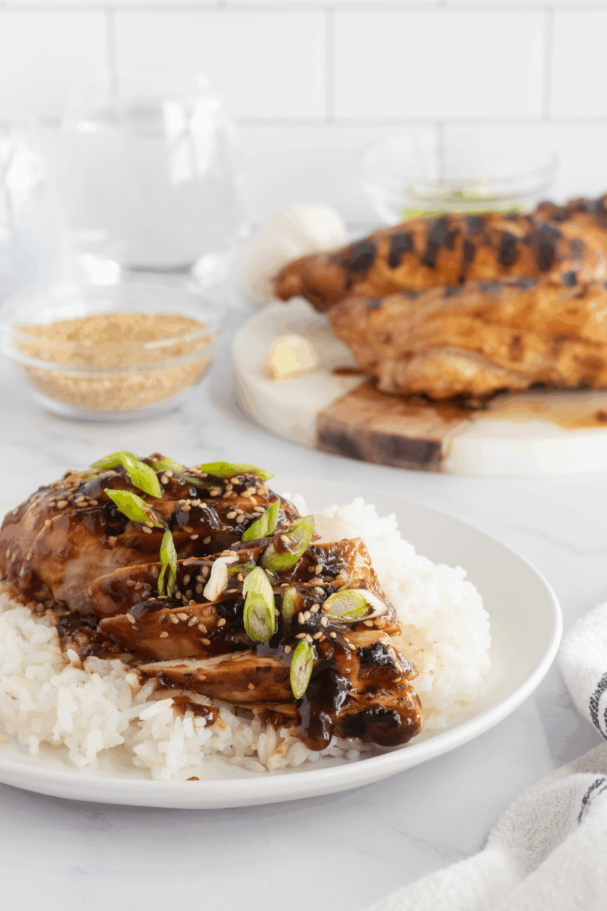 A plate of white rice topped with glazed teriyaki chicken, sliced green onions, and sesame seeds. In the background, more chicken, a bowl of sesame seeds, and glassware are visible on a white countertop.