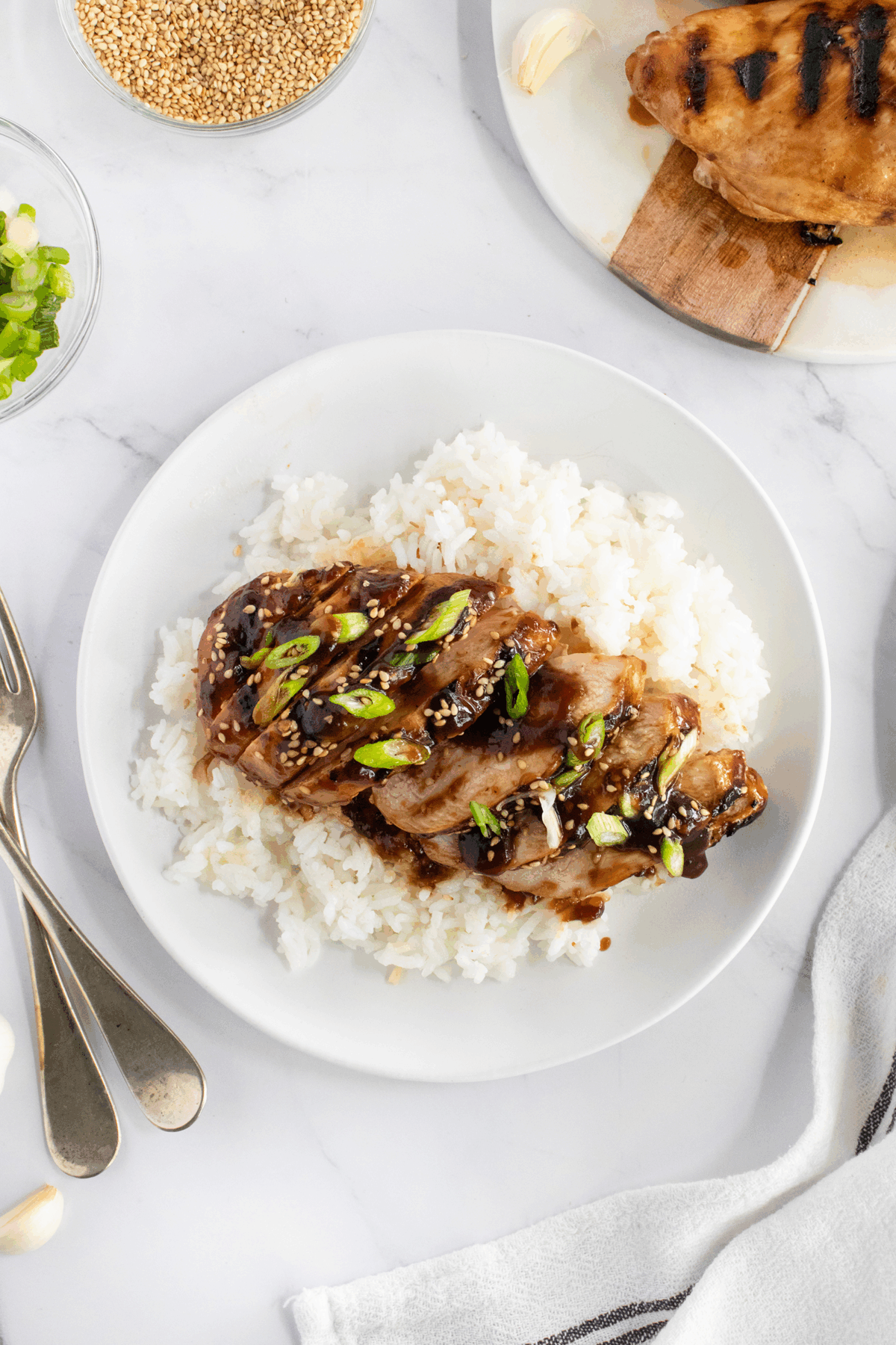 A white plate with grilled chicken slices glazed in sauce, topped with chopped green onions and sesame seeds, served over a bed of white rice. Utensils and side dishes are visible around the plate on a marble surface.