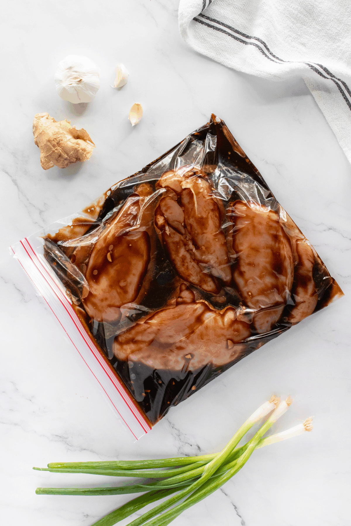 A zip-top bag containing raw chicken marinating in a dark sauce is placed on a marble countertop. Nearby are garlic cloves, a piece of ginger, green onions, and part of a white cloth with black stripes.