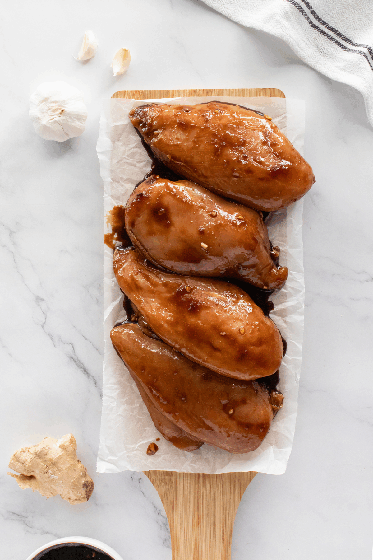 Four glazed chicken breasts arranged on a parchment-lined wooden board, with whole garlic, a piece of ginger, and a small bowl of dark sauce nearby on a marble surface.