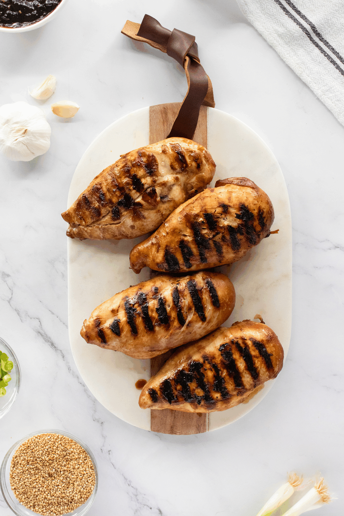 Four grilled chicken breasts with char marks are arranged on a marble cutting board with a wooden handle. Surrounding the board are bowls of mustard seeds, green onions, whole garlic, and a kitchen towel.