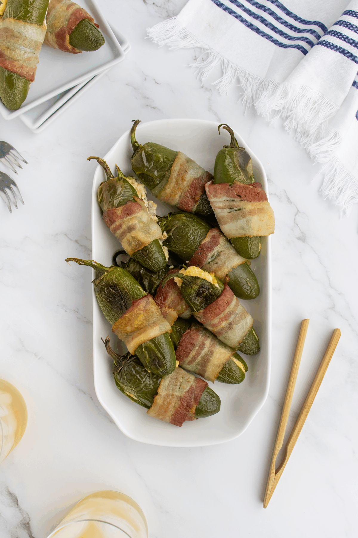 A white platter holds several bacon-wrapped jalapeño poppers filled with cheese, placed on a marble surface. Wooden chopsticks and drinks are nearby, with a blue-striped napkin in the background.
