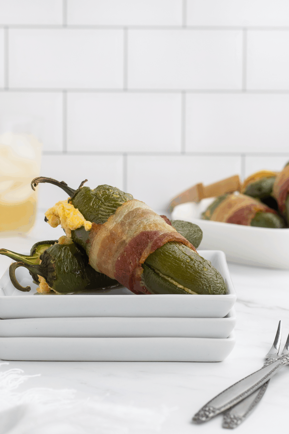 Three bacon-wrapped jalapeño poppers stuffed with cheese are stacked on a small white plate, with more poppers blurred in the background against a white tiled wall.