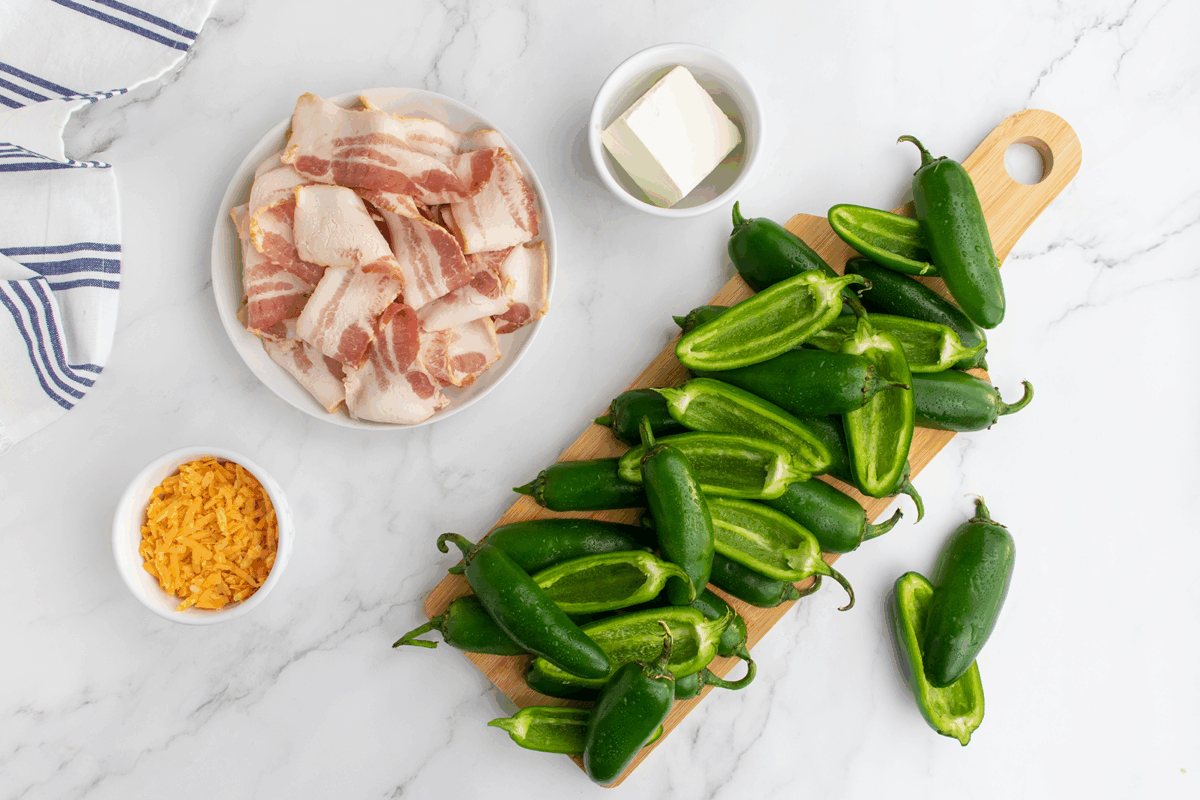 A cutting board with halved jalapeños, next to bowls of shredded cheddar, cream cheese, and a plate of raw bacon slices on a white marble surface.
