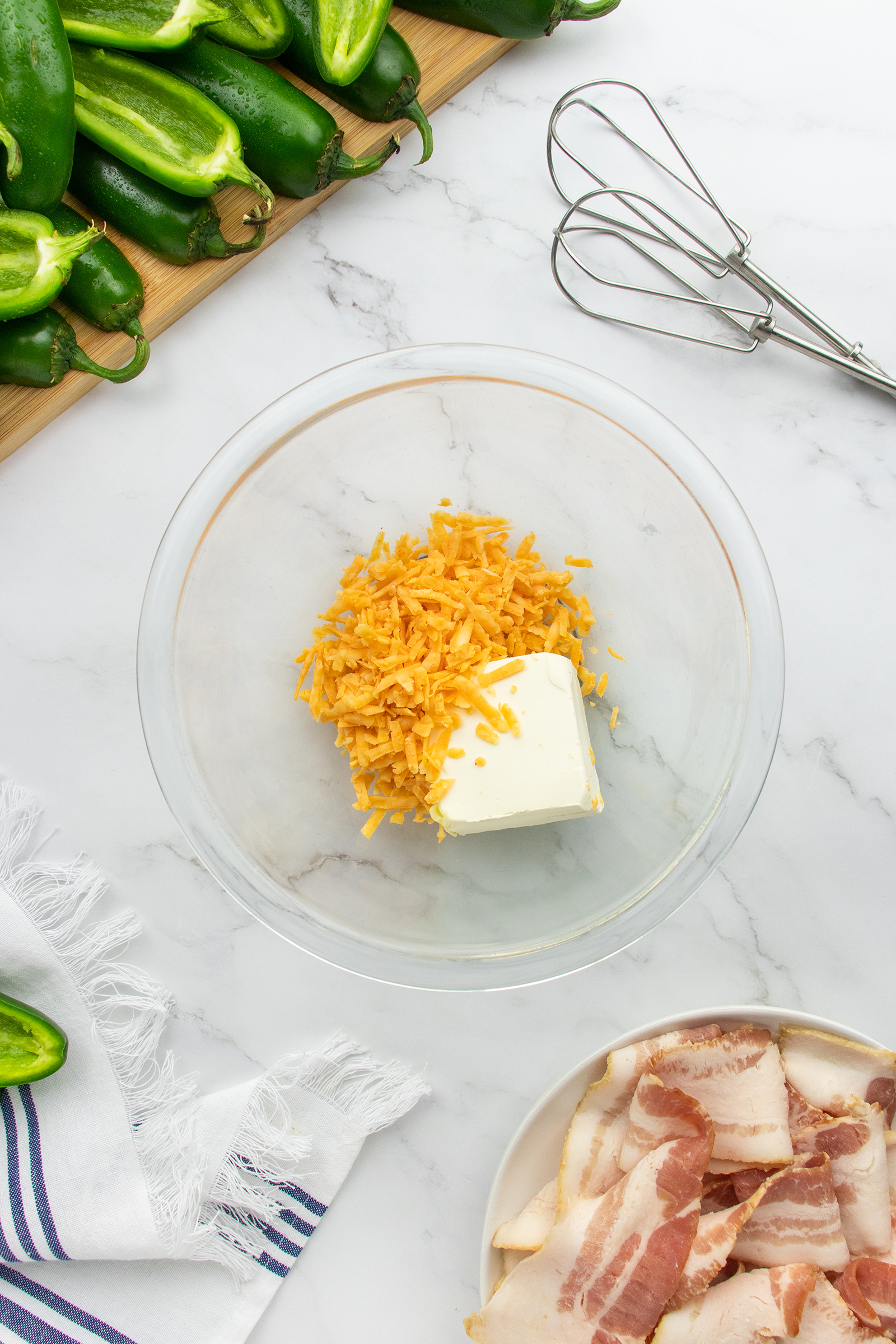 A glass bowl with shredded cheddar cheese and cream cheese sits on a marble countertop, surrounded by jalapeños, a whisk, bacon slices on a plate, and a striped towel.