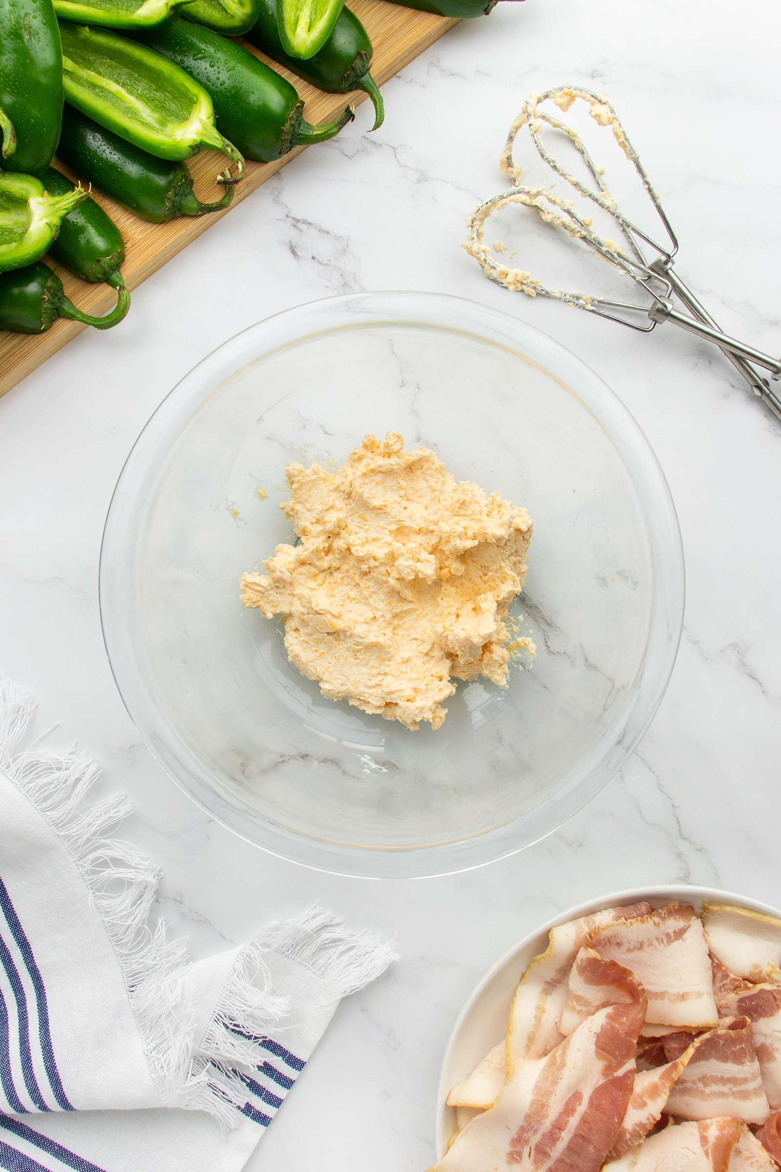 A glass bowl with a creamy cheese mixture sits on a marble surface beside sliced jalapeños, raw bacon on a plate, a hand mixer with beaters, and a white cloth with blue stripes.