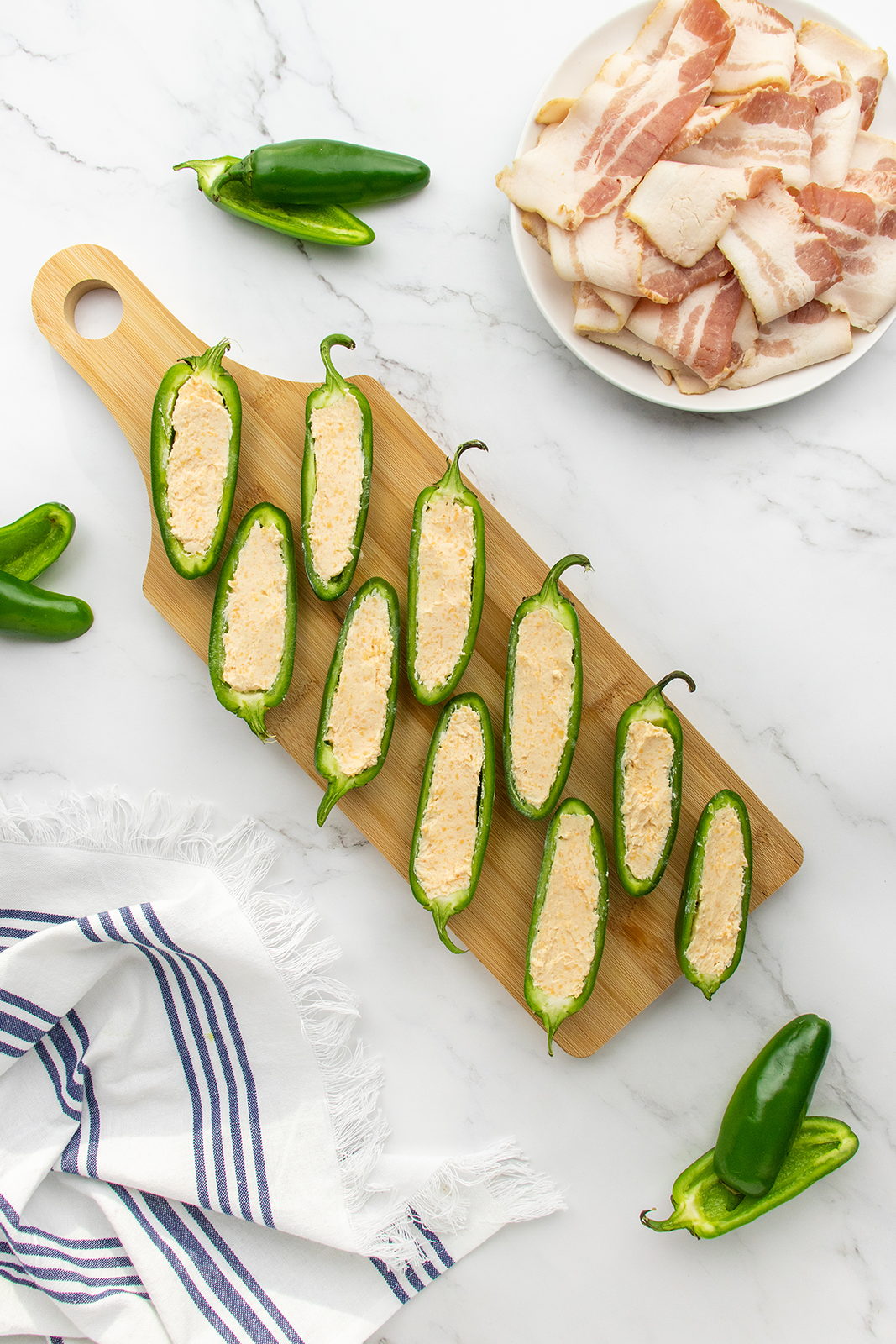 A wooden board holds halved jalapeños filled with creamy cheese mixture. Nearby are whole jalapeños, a bowl of raw bacon slices, and a striped kitchen towel on a white marble surface.