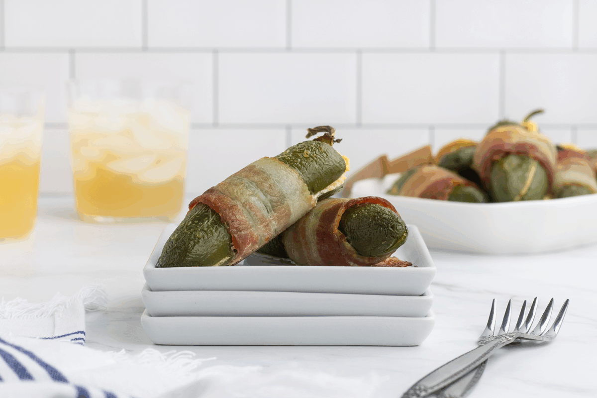 Three bacon-wrapped jalapeño poppers stacked on small white plates, with more poppers in the background and two iced drinks on a white marble surface against a white tiled wall.