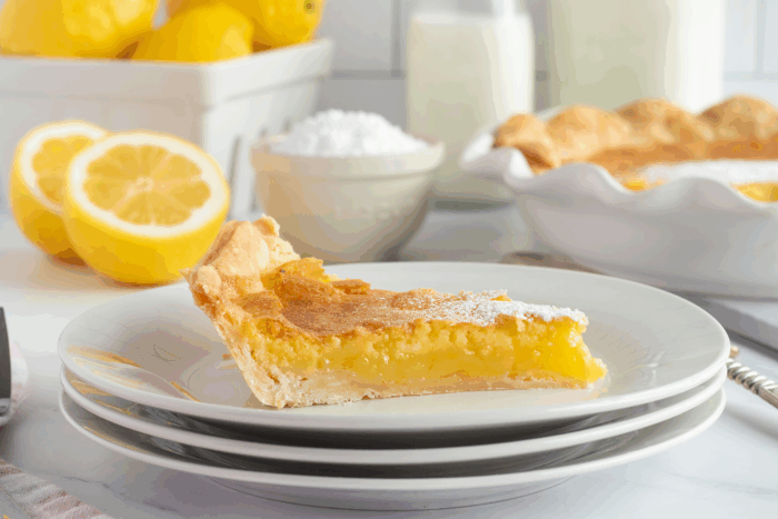 A slice of lemon pie on a stack of white plates, with fresh lemons, a bowl of powdered sugar, a glass of milk, and a whole pie in the background.