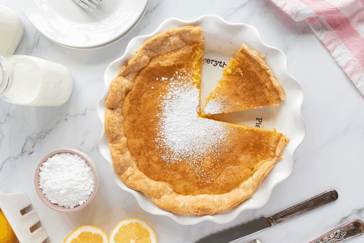 A golden pie with a flaky crust, partially sliced, sits in a white dish. The pie is dusted with powdered sugar. Nearby are lemon slices, a small bowl of powdered sugar, a bottle of milk, and a knife on a marble surface.