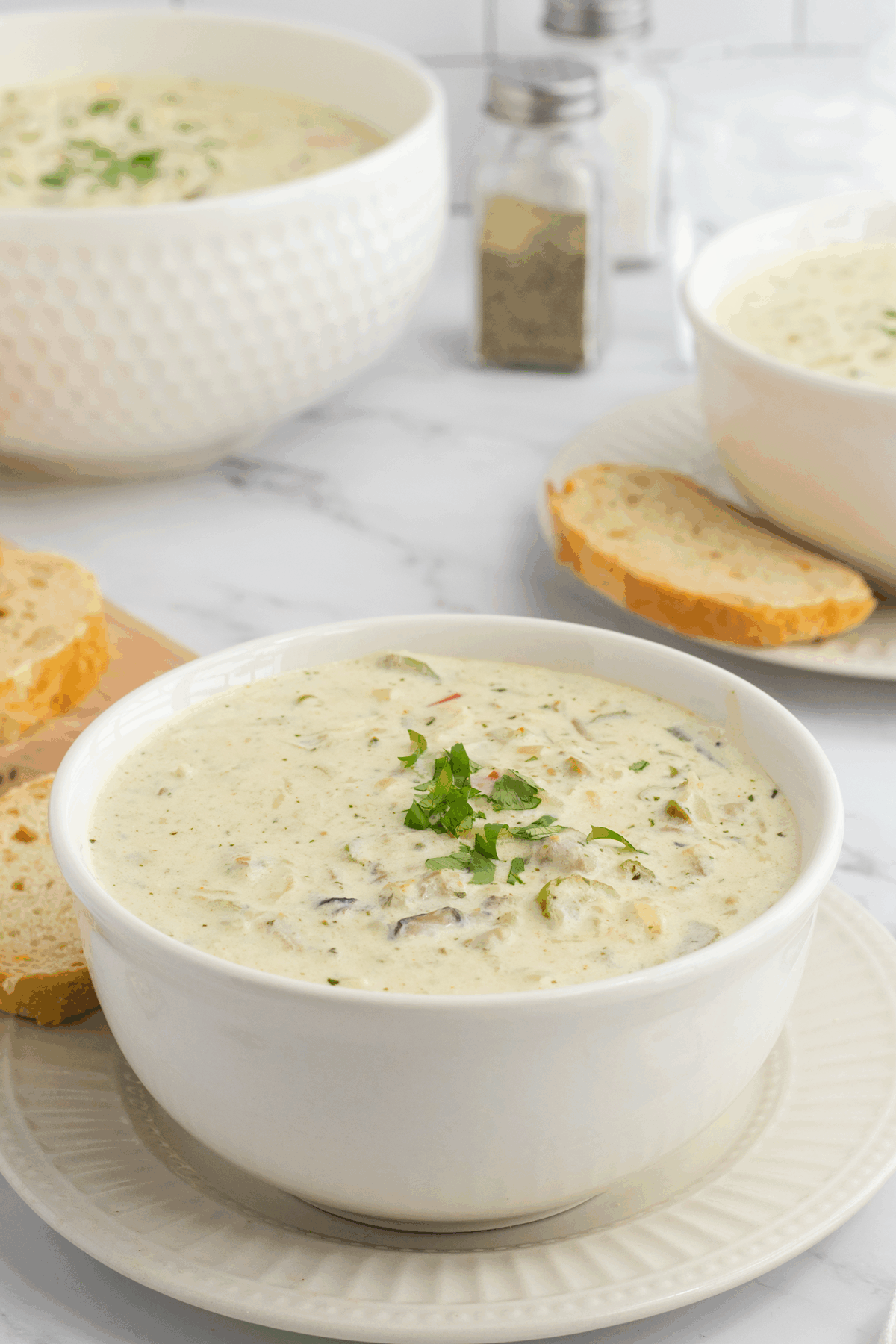 A white bowl filled with creamy soup, garnished with chopped herbs, sits on a plate next to slices of bread. Another bowl of soup, a serving dish, and salt and pepper shakers are in the background.