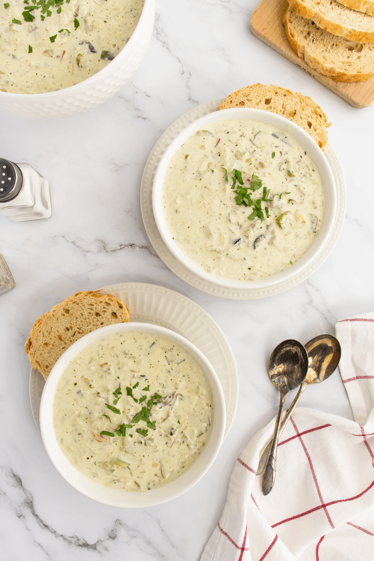 Two bowls of creamy soup garnished with chopped herbs, each served with a slice of bread on white plates. Spoons and a red-striped napkin are nearby on a marble surface; sliced bread is visible in the background.