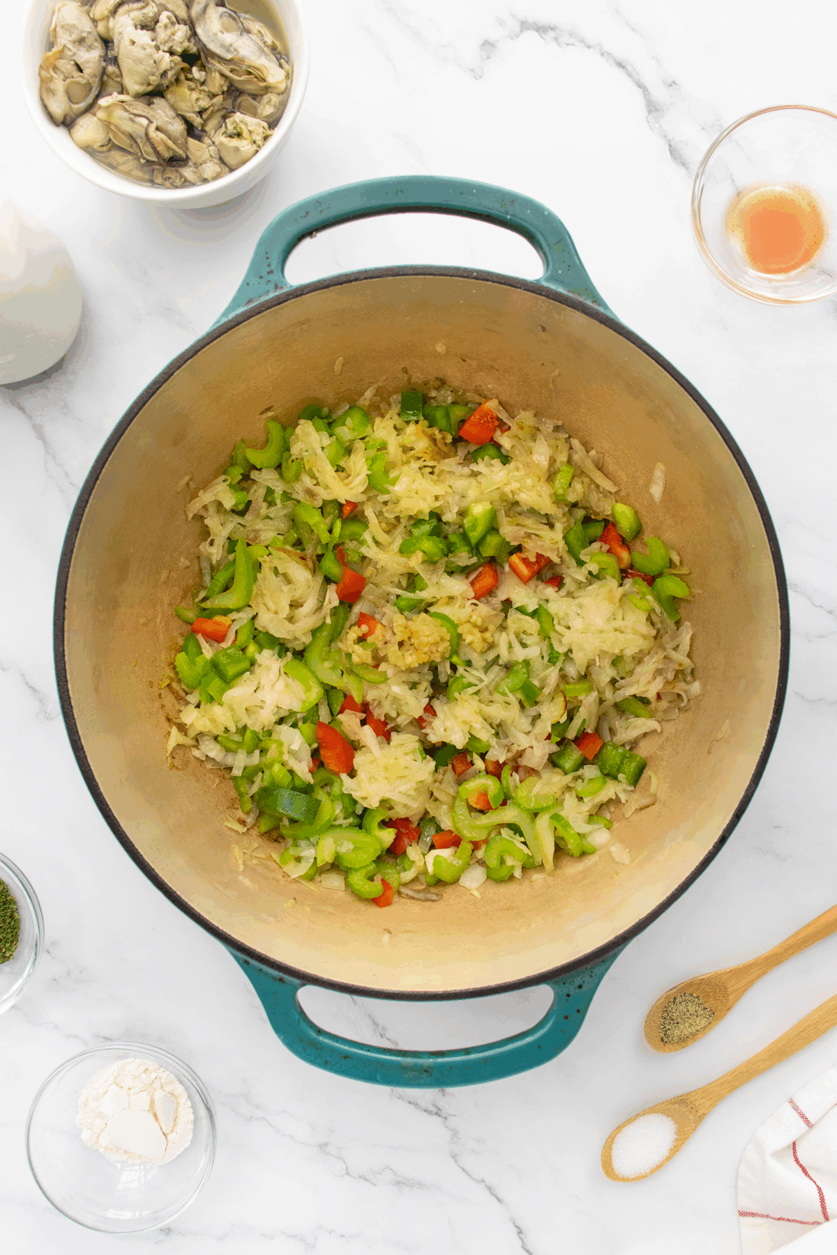 A large pot on a marble counter contains sautéed chopped onions, green bell peppers, and red bell peppers. Surrounding the pot are small bowls with seasonings, flour, a bowl of oysters, and a cup of liquid.