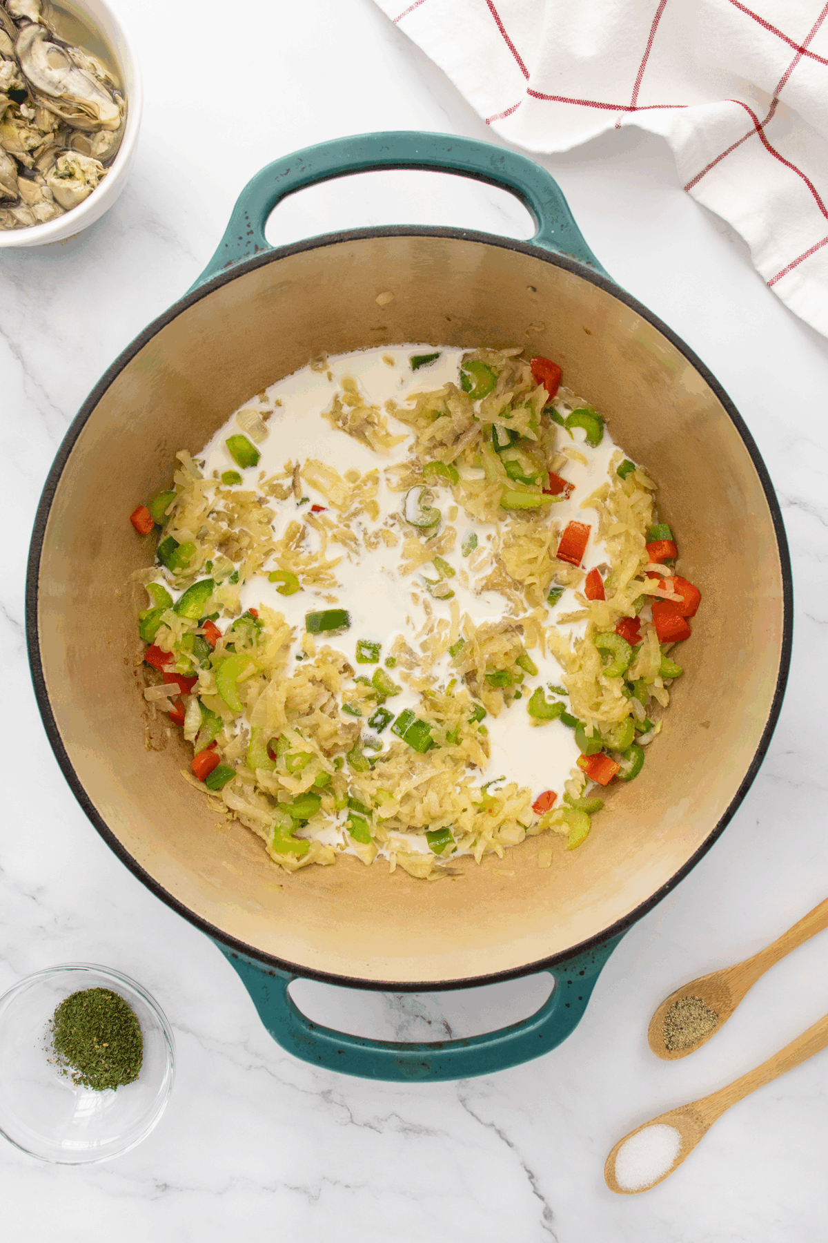 A large pot on a marble countertop holds chopped onions, celery, green and red bell peppers, and cream. Nearby are bowls with oysters, dried herbs, and a striped towel, plus wooden spoons.