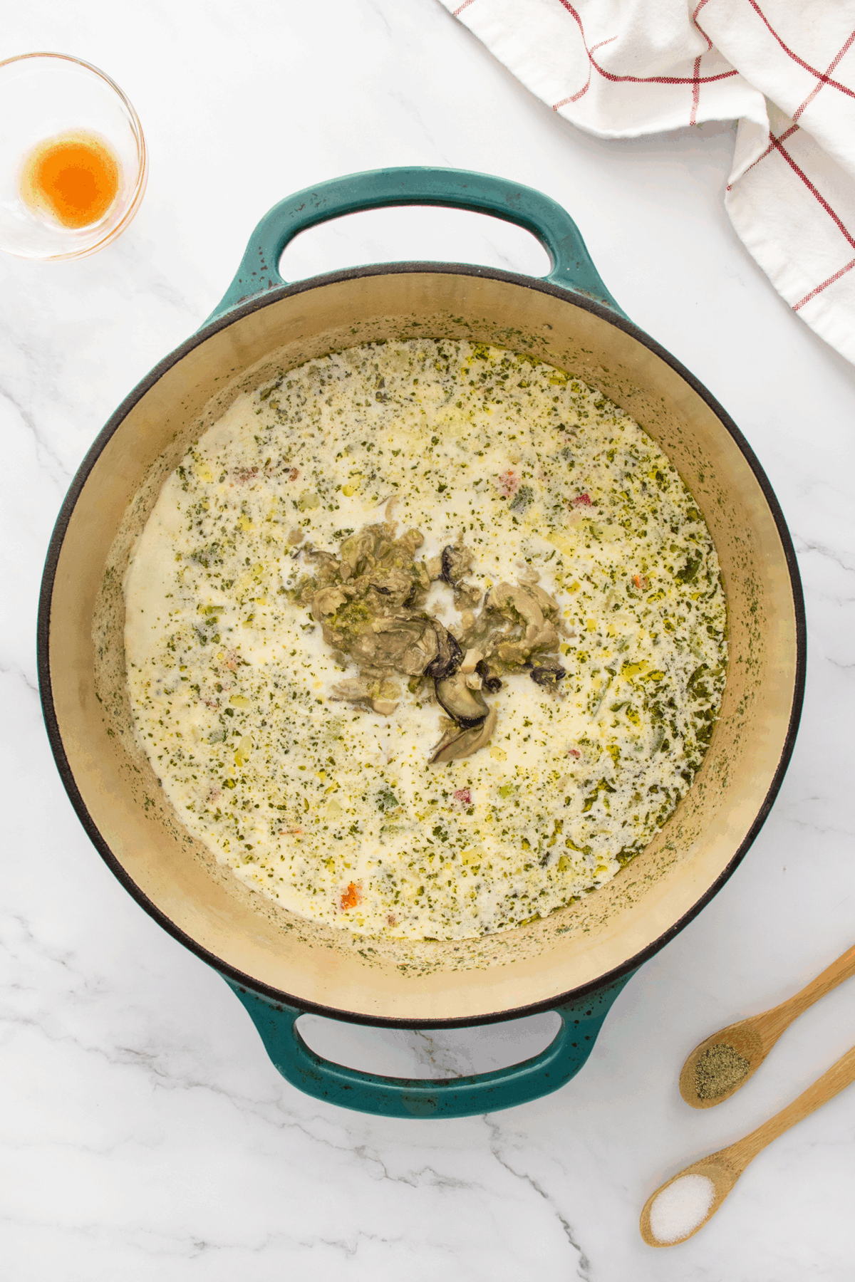 A large teal Dutch oven filled with creamy soup, herbs, and mushrooms sits on a marble countertop. Nearby are a wooden spoon, a measuring spoon with pepper, a glass bowl, and a white cloth with red stripes.