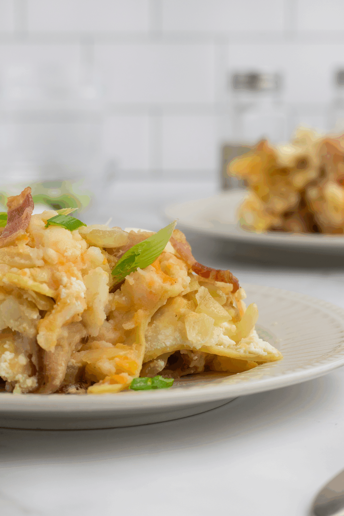 A close-up of a serving of cheesy potato casserole topped with green onions on a plate, with another plate of casserole and some seasoning jars blurred in the background.