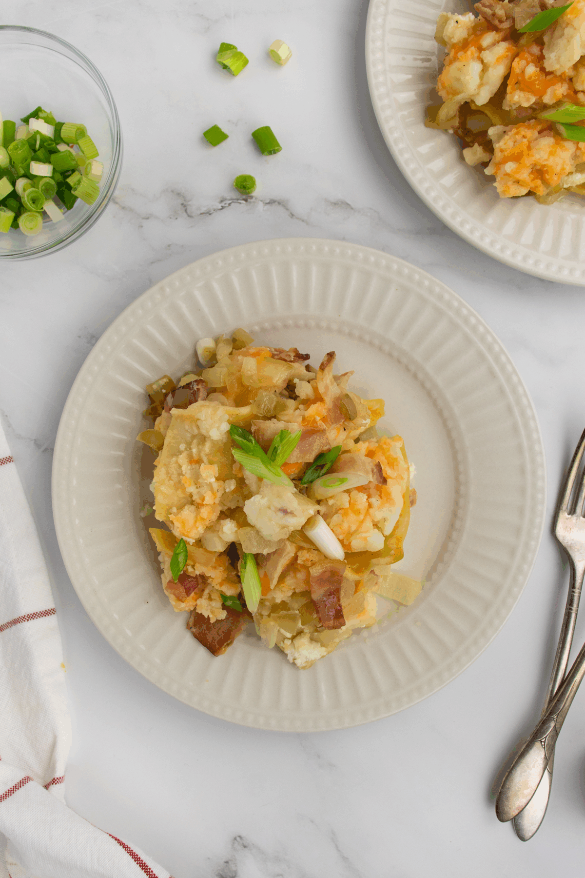 A serving of a casserole with melted cheese, potatoes, onions, and green onions is plated on a white dish. A fork, napkin, and small bowl of chopped green onions are nearby on a marble surface.