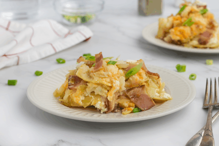 A serving of cheesy potato casserole topped with chopped green onions on a white plate, with a fork beside it. Another plate of casserole and kitchen items are blurred in the background.