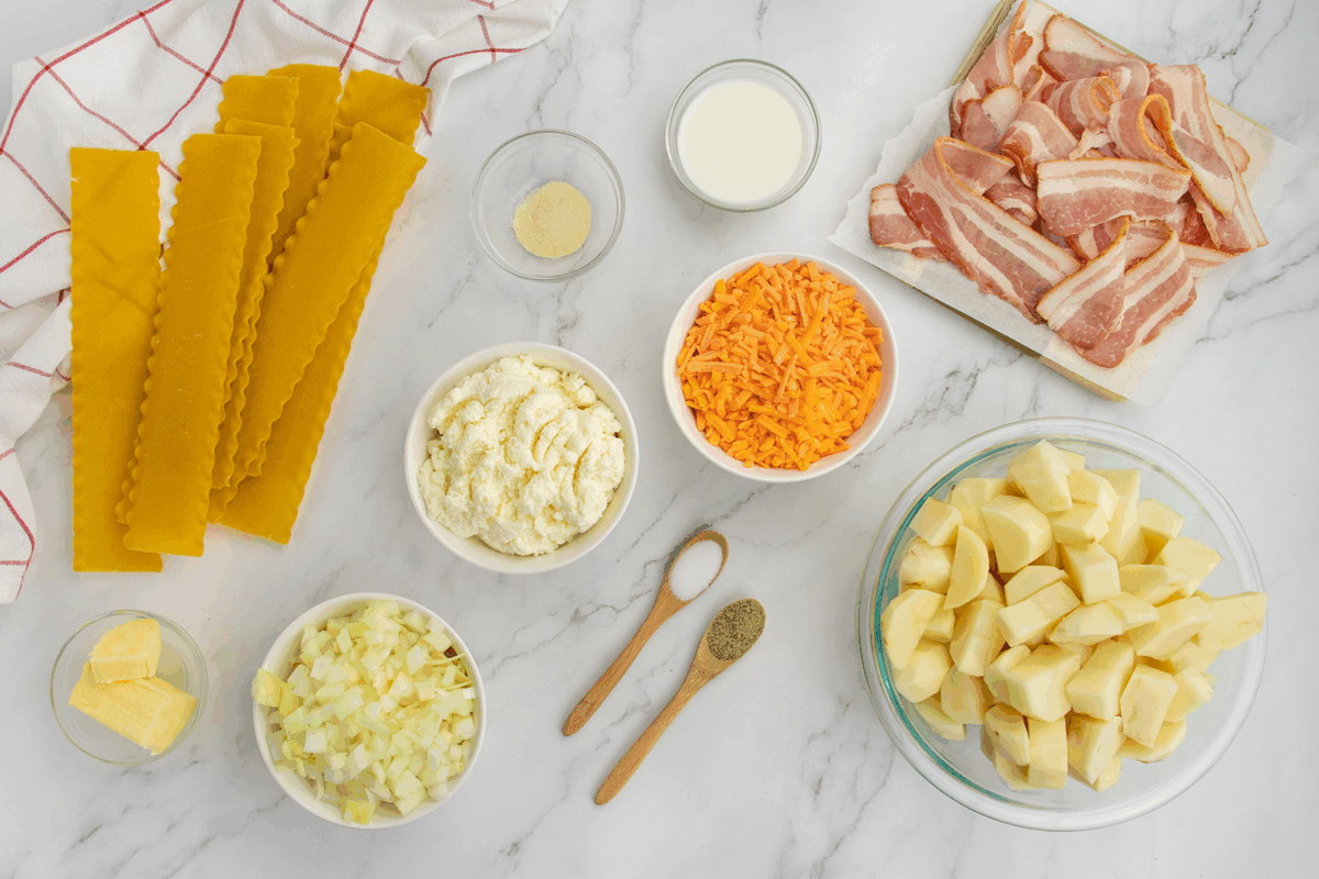 Lasagna noodles, diced potatoes, shredded cheddar, ricotta, chopped onion, bacon slices, butter, milk, granulated onion, salt, and pepper are arranged on a marble surface with a kitchen towel in the background.