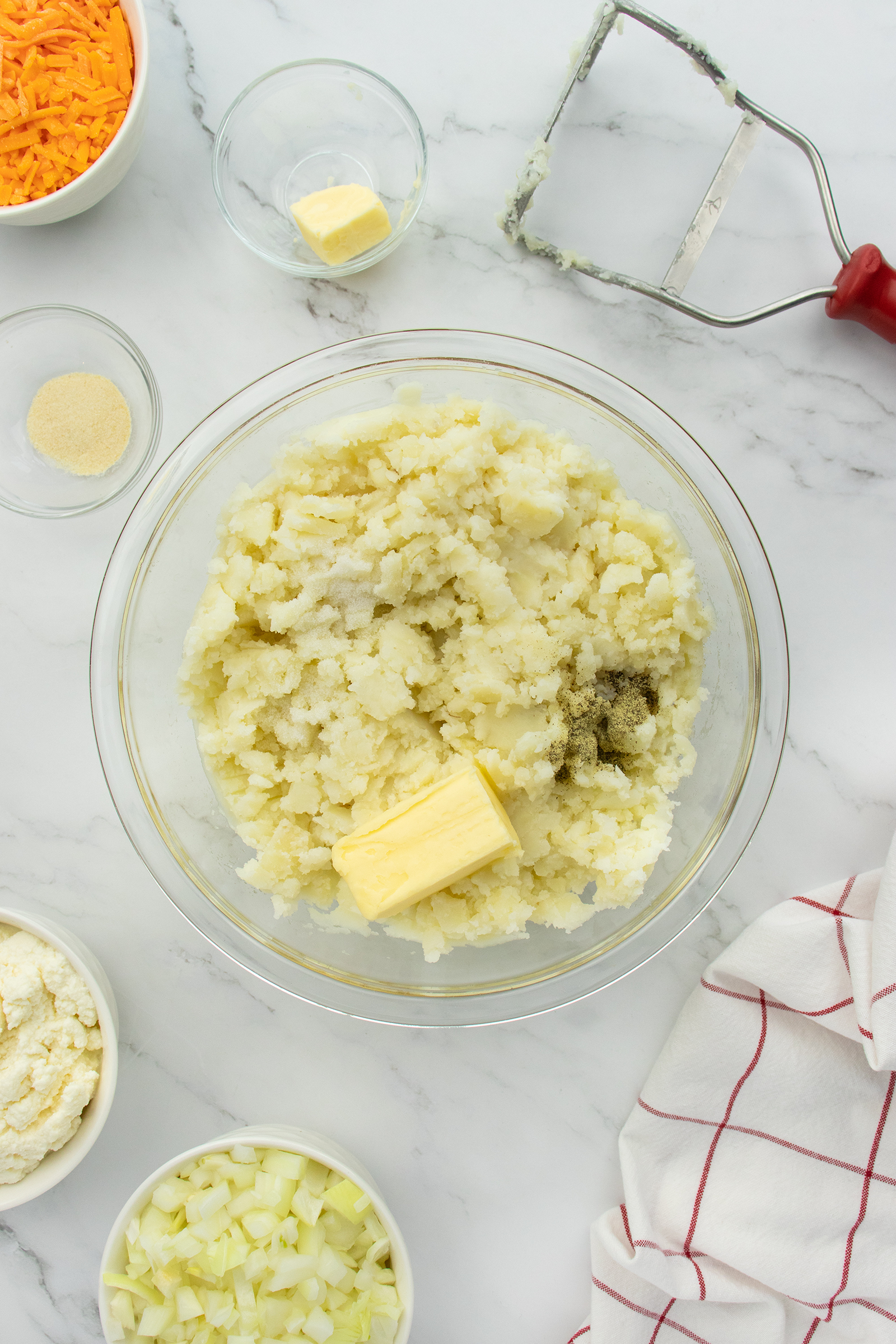 A glass bowl with mashed potatoes, a stick of butter, and seasoning on a marble counter, surrounded by chopped onions, shredded cheese, a potato masher, and a red-striped kitchen towel.
