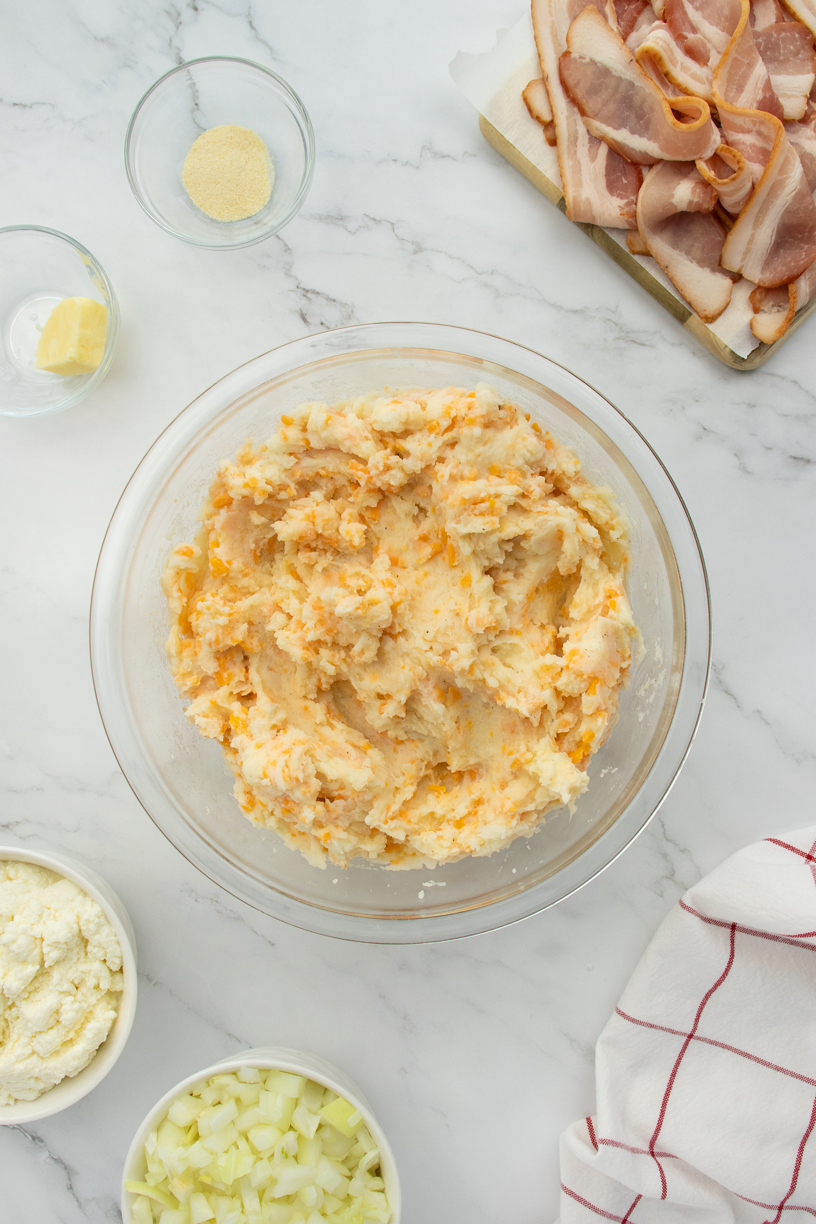 A bowl of mashed potatoes mixed with cheese sits on a marble countertop. Surrounding it are bowls of chopped onions, cheese, seasoning, a pat of butter, and a tray of raw bacon. A red-striped towel is at the side.