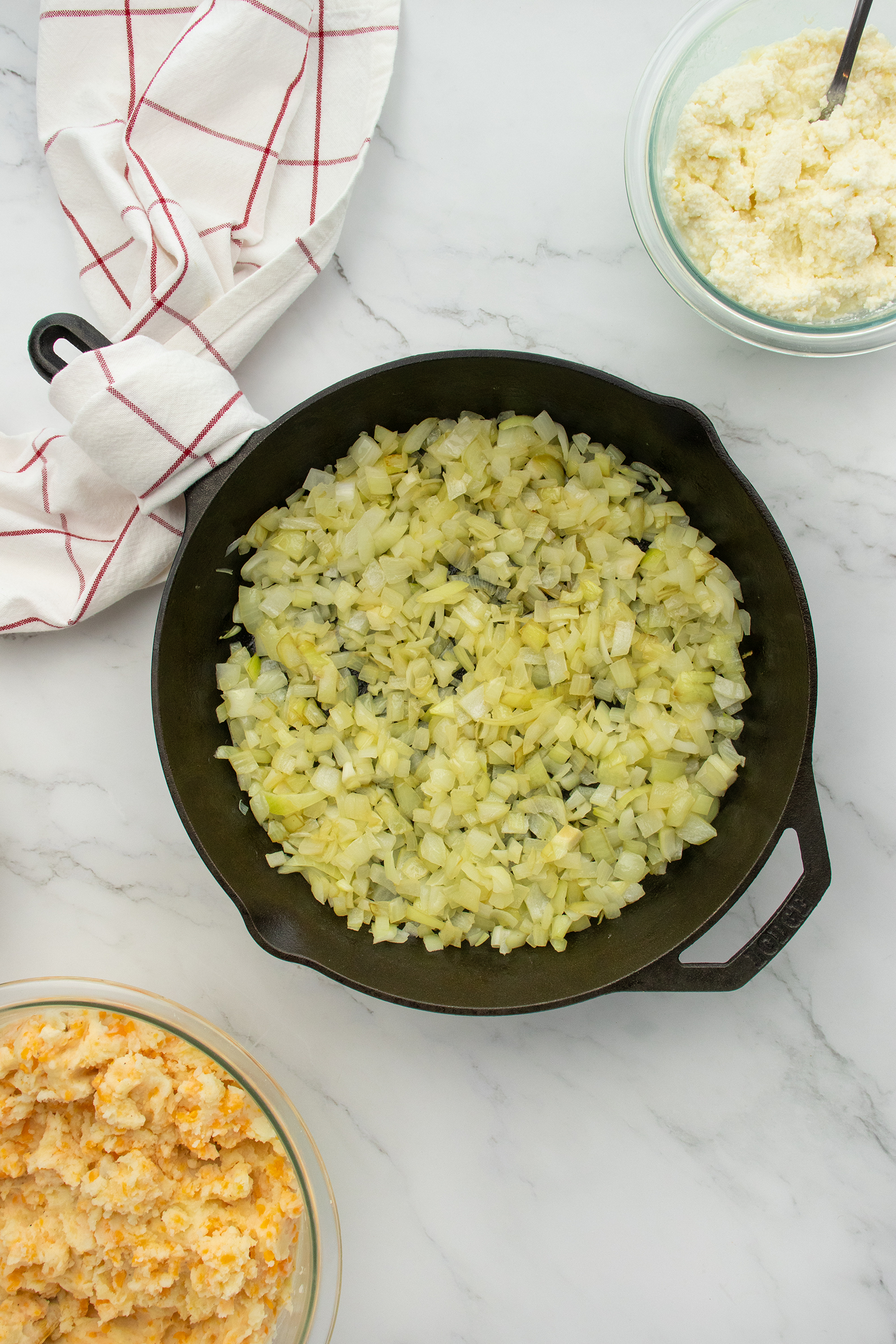 A cast iron skillet filled with chopped, cooked onions sits on a marble countertop. Nearby are a bowl of ricotta cheese, a bowl of orange-tinted cheese mixture, and a white towel with red stripes.