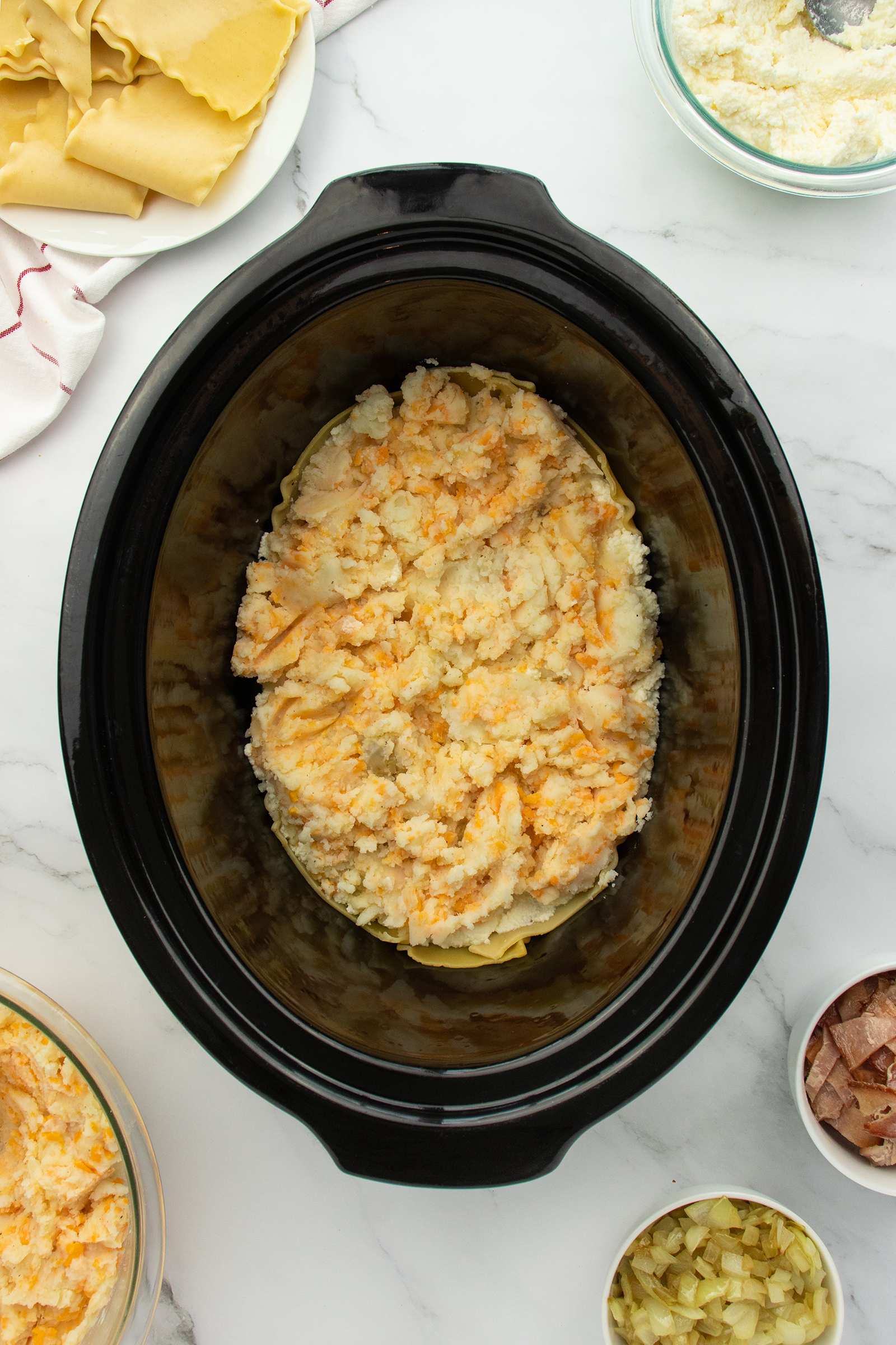 A slow cooker filled with layered uncooked lasagna, topped with a cheese mixture. Surrounding the slow cooker are bowls with ricotta, sautéed onions, and chopped ham, and a plate with uncooked lasagna noodles.