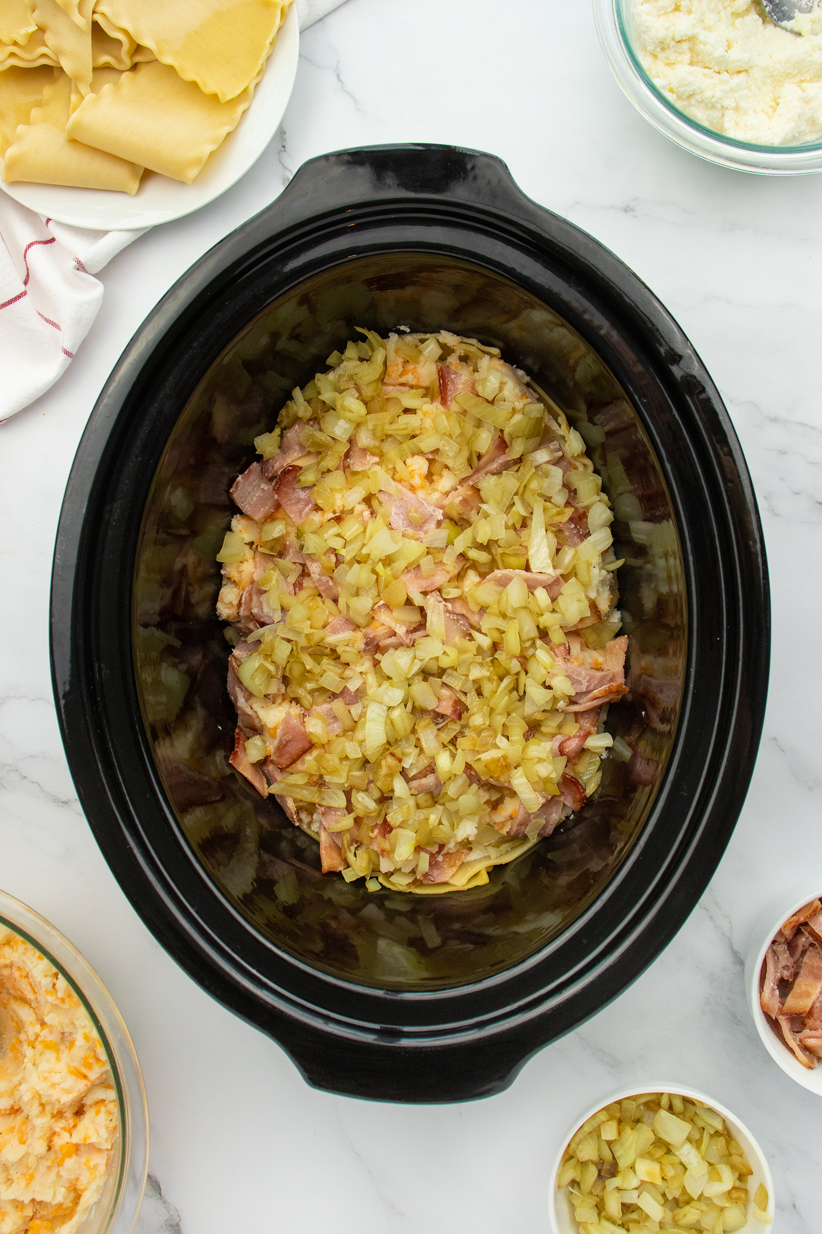 A black slow cooker filled with chopped cooked bacon and onions sits on a white surface, surrounded by bowls of cheese, ricotta, chopped pickles, and uncooked lasagna noodles.