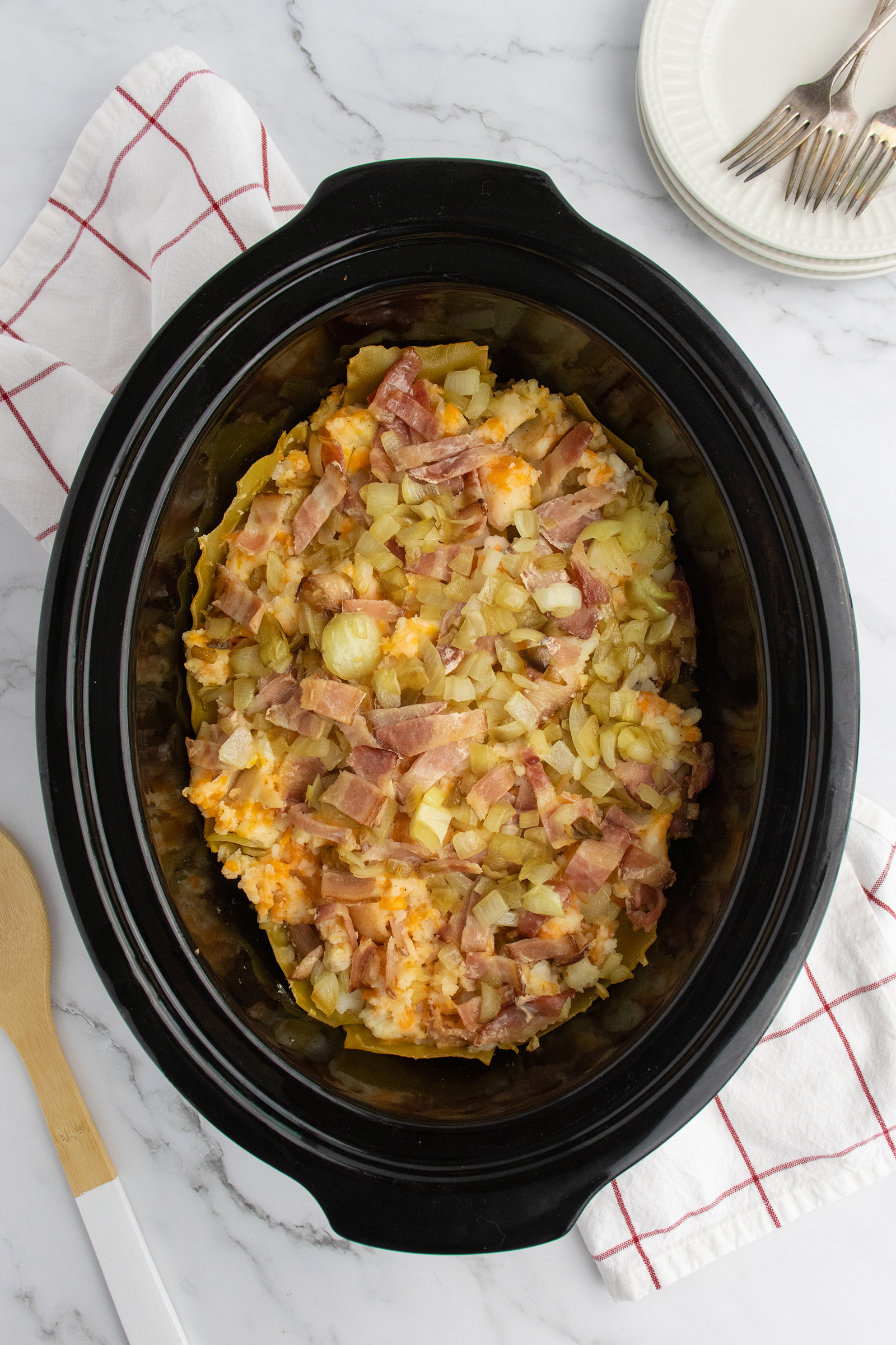 A slow cooker filled with a cheesy potato casserole topped with chopped onions and bacon sits on a marble countertop. Nearby are a folded white cloth with red stripes, a wooden spoon, and stacked plates with forks.