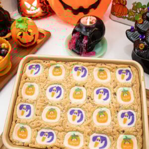 A baking tray of cookie bars topped with Halloween-themed cookies featuring pumpkins and ghosts sits on a wooden board, surrounded by Halloween decorations like jack-o-lanterns and pumpkins.