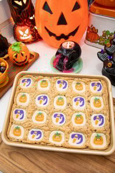 A baking tray of cookie bars topped with Halloween-themed cookies featuring pumpkins and ghosts sits on a wooden board, surrounded by Halloween decorations like jack-o-lanterns and pumpkins.