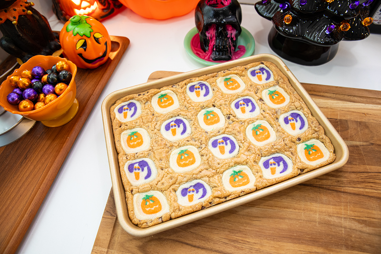 A rectangular baking pan with Halloween-themed cookie bars, featuring ghost and pumpkin designs, sits on a wooden board. Festive Halloween decorations surround it, including pumpkins, a black skull, and a tree.