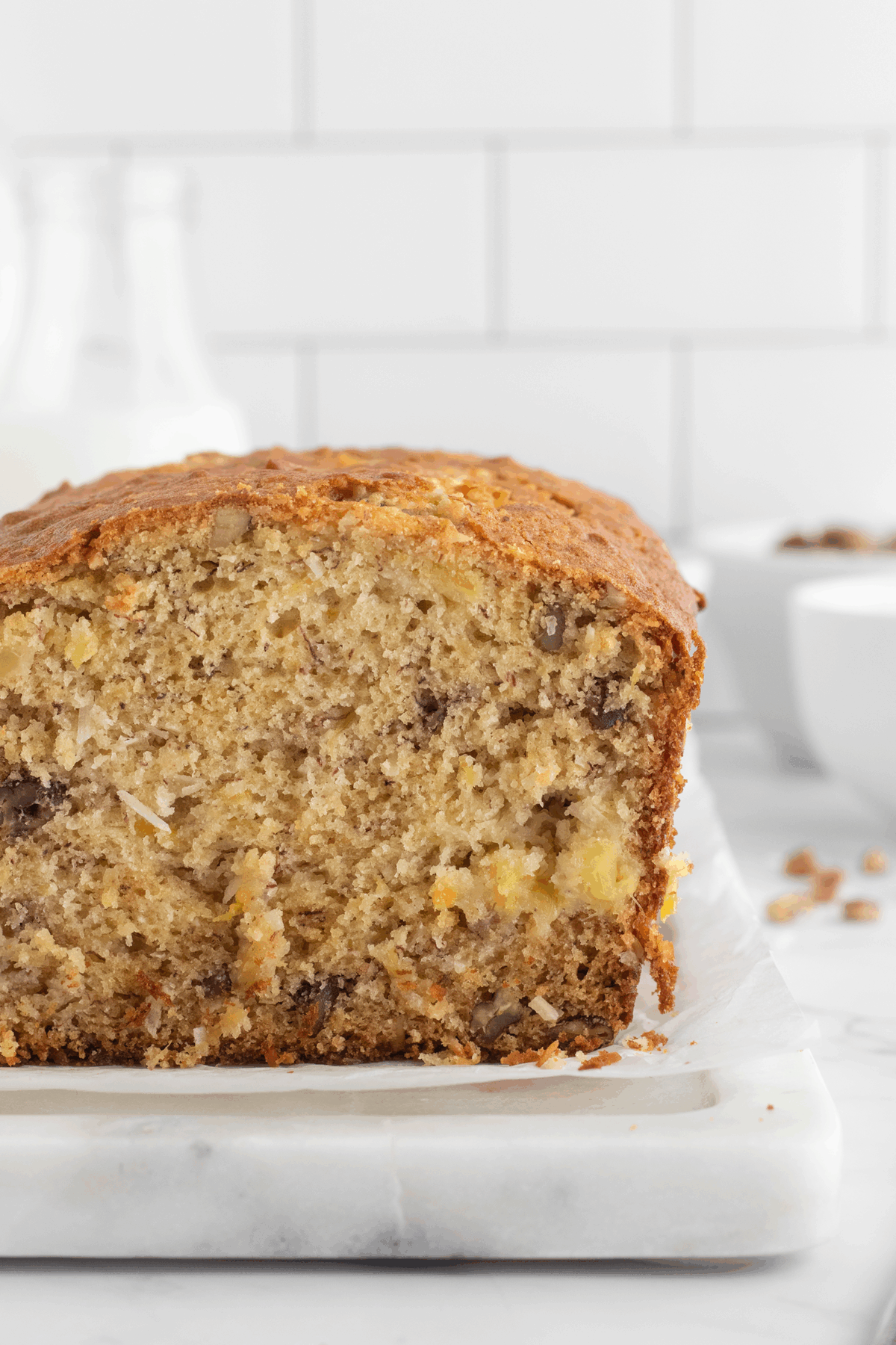 A close-up of a sliced loaf of banana bread with visible nuts on a marble surface, set against a white tiled background.
