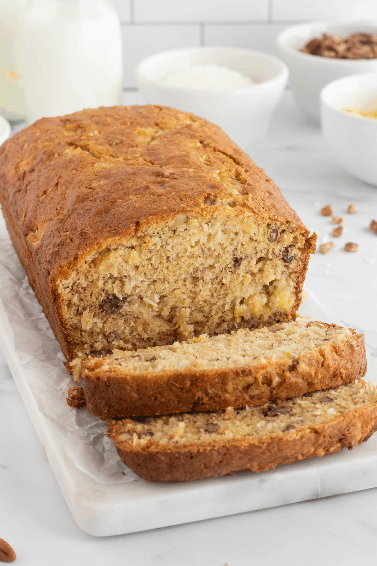 A loaf of banana bread with a golden crust sits on a white marble board. Two slices are cut in front, showing a moist, textured interior with bits of walnut. Blurry bowls and a milk bottle are in the background.