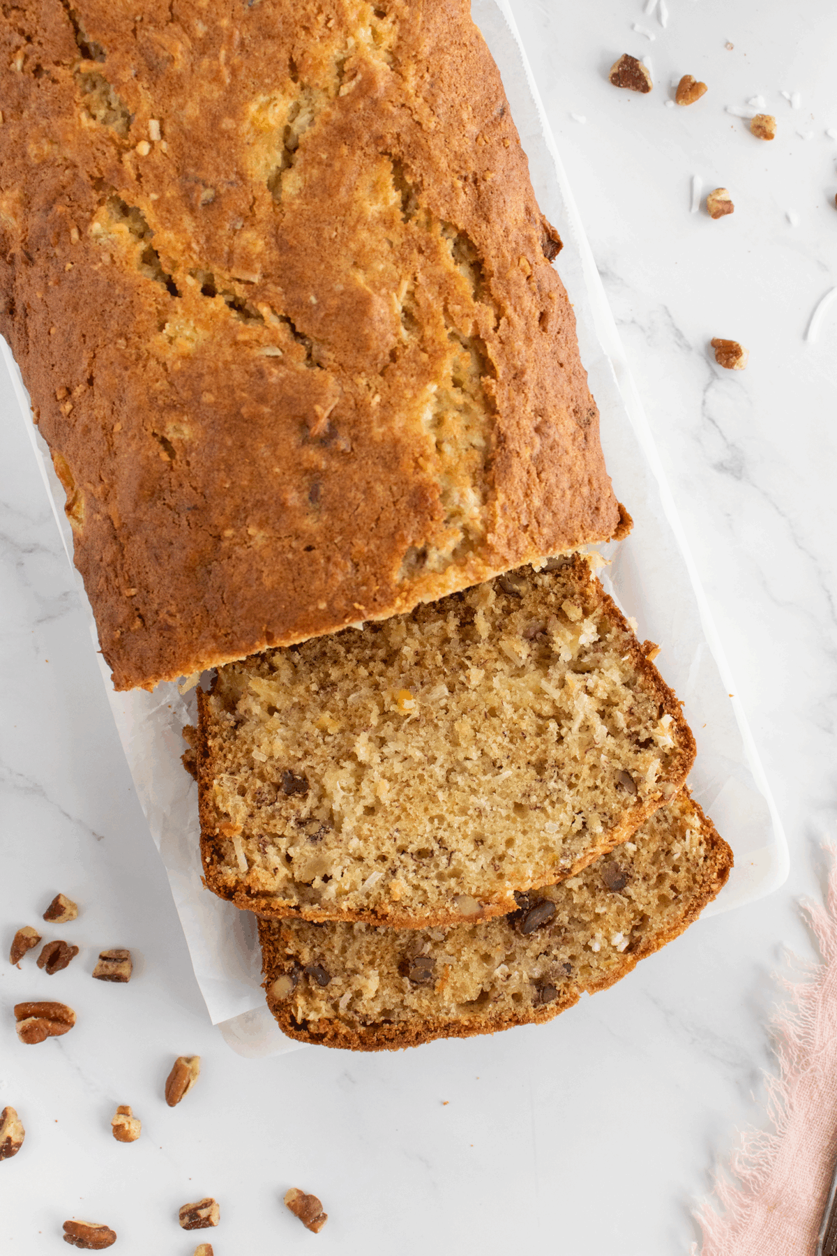 A loaf of banana bread on a white surface with two slices cut, showing a moist texture with pieces of walnut or pecan inside. Small nut pieces are scattered around the loaf.