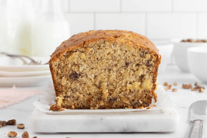 A sliced loaf of banana bread with visible nuts on a piece of parchment paper, placed on a marble board. The background includes blurred plates, milk bottles, and bowls on a white countertop.
