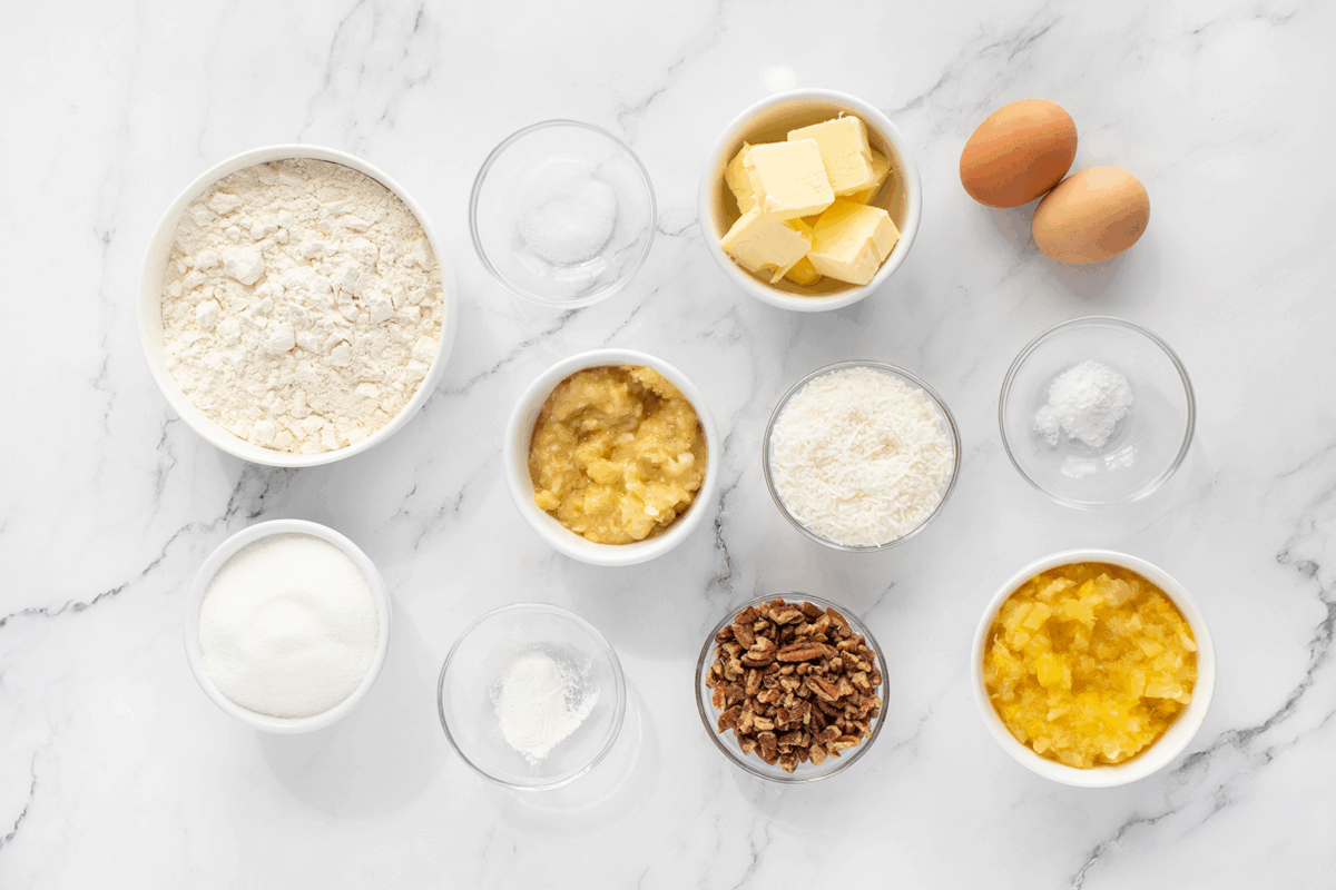 Various baking ingredients arranged on a white marble surface, including flour, sugar, eggs, butter, chopped pecans, crushed pineapple, mashed bananas, shredded coconut, baking powder, and salt in small bowls.