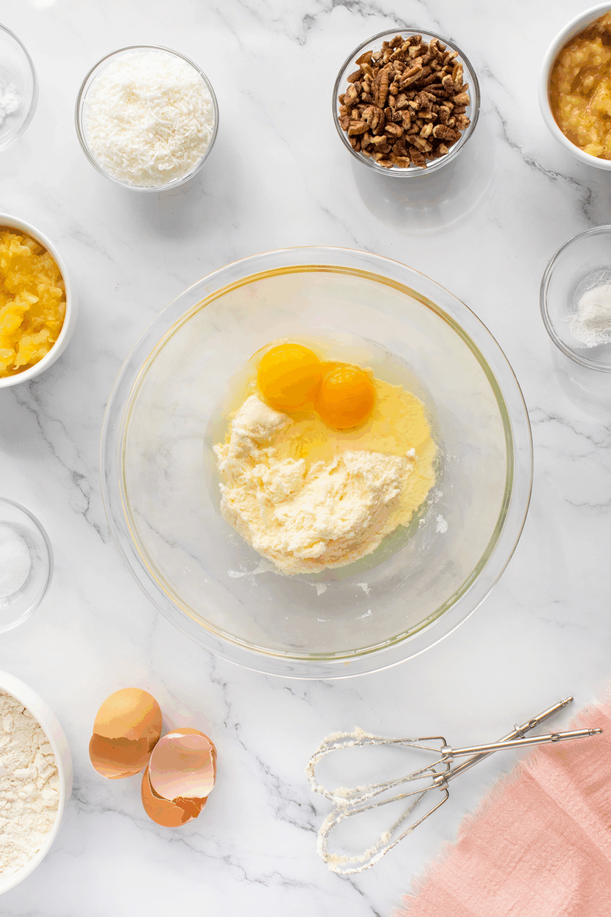 A glass bowl with eggs and flour mixture sits on a marble countertop, surrounded by bowls of chopped pecans, shredded coconut, pineapple, mashed banana, flour, and an egg beater beside egg shells and a pink cloth.
