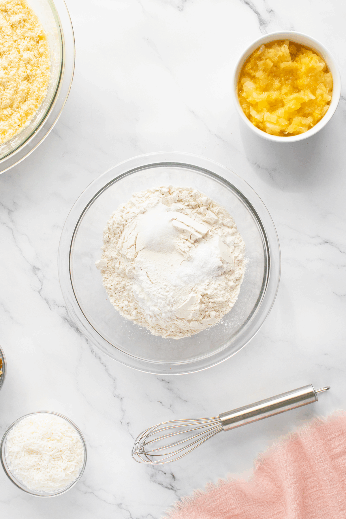 A glass bowl of flour mixture sits on a marble countertop surrounded by a whisk, a bowl of crushed pineapple, shredded coconut, and other baking ingredients. A pink cloth is partially visible in the corner.