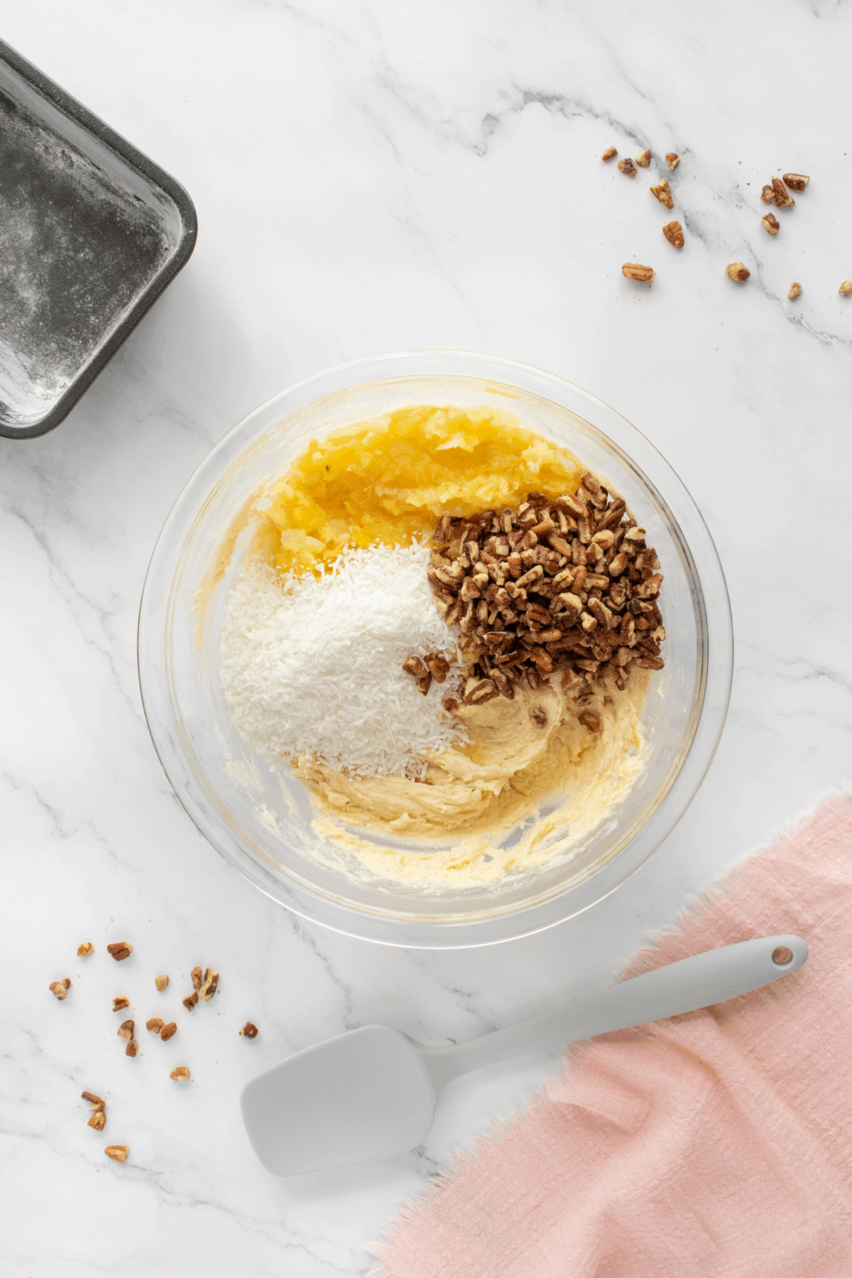 A glass bowl with cake batter, shredded coconut, crushed pineapple, and chopped pecans on a marble surface. Nearby are a spatula, a loaf pan, and scattered pecans. A pink cloth lies to the side.