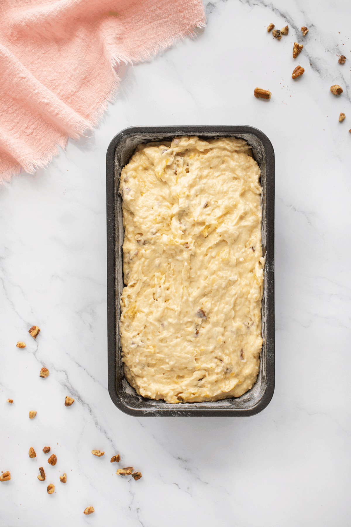 A loaf pan filled with unbaked banana bread batter sits on a white marble surface, surrounded by scattered pecans and a pink cloth in the corner.