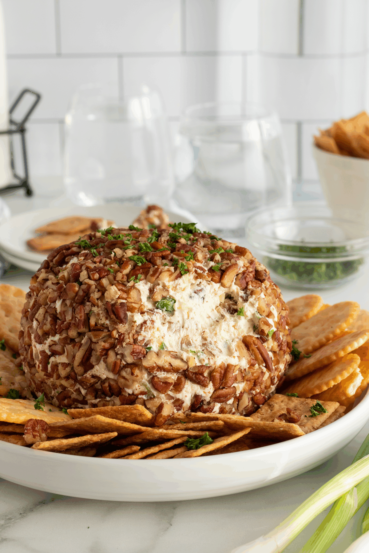 A round cheese ball coated in chopped pecans sits on a white plate, surrounded by rectangular crackers. Chopped herbs are sprinkled on top, and there are glasses, plates, and a bowl of crackers in the background.