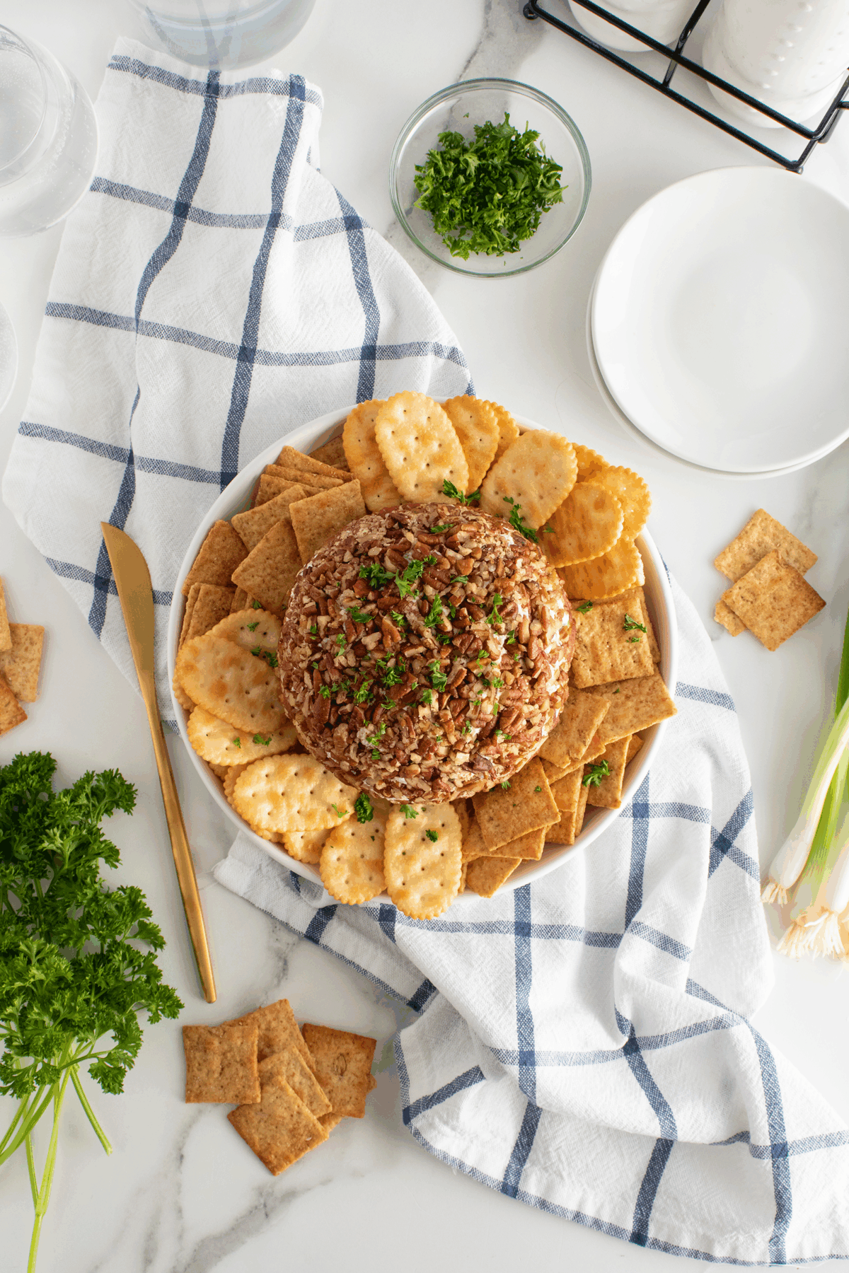 A round cheese ball topped with chopped pecans and herbs is surrounded by assorted crackers on a white plate, set on a blue and white checkered towel with parsley, green onions, and plates nearby.
