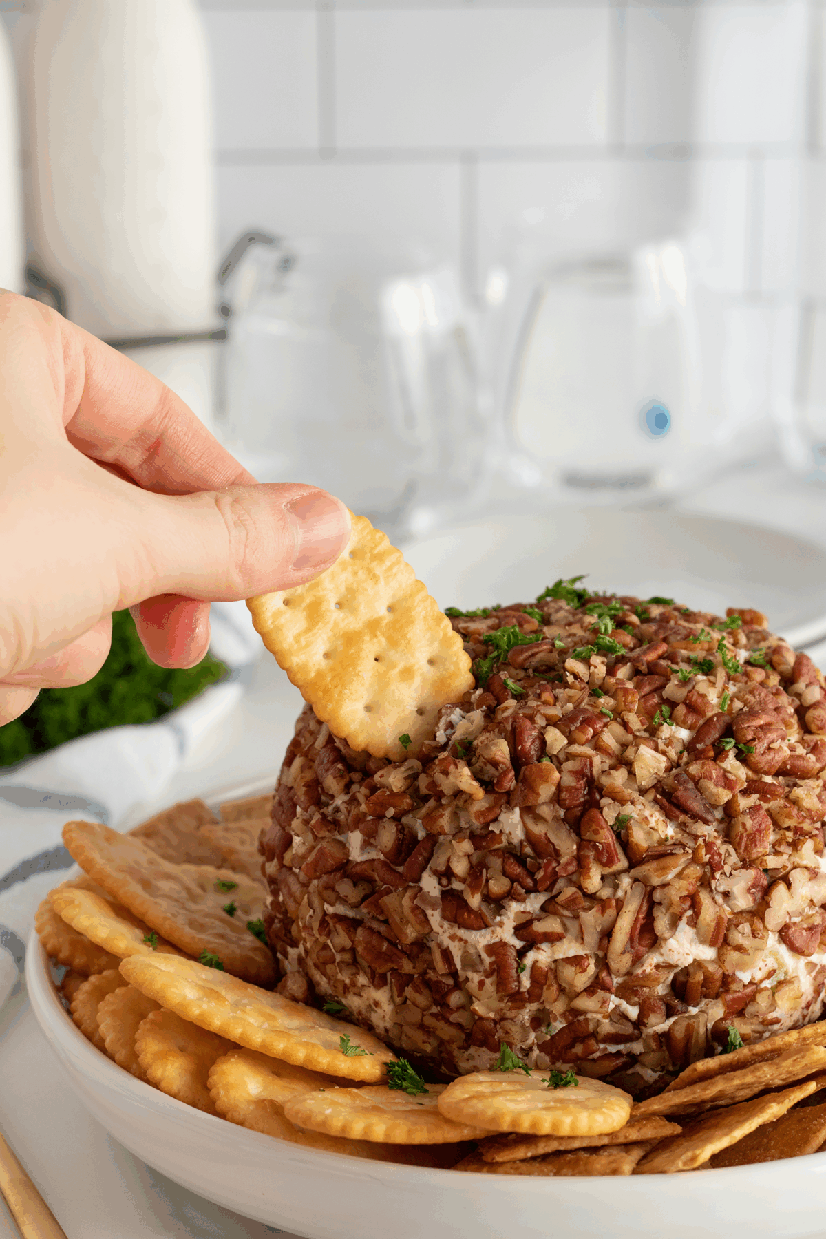 A hand dips a square cracker into a round cheese ball coated with chopped pecans, served on a white plate with assorted crackers. The background shows glasses and a milk bottle on a white countertop.