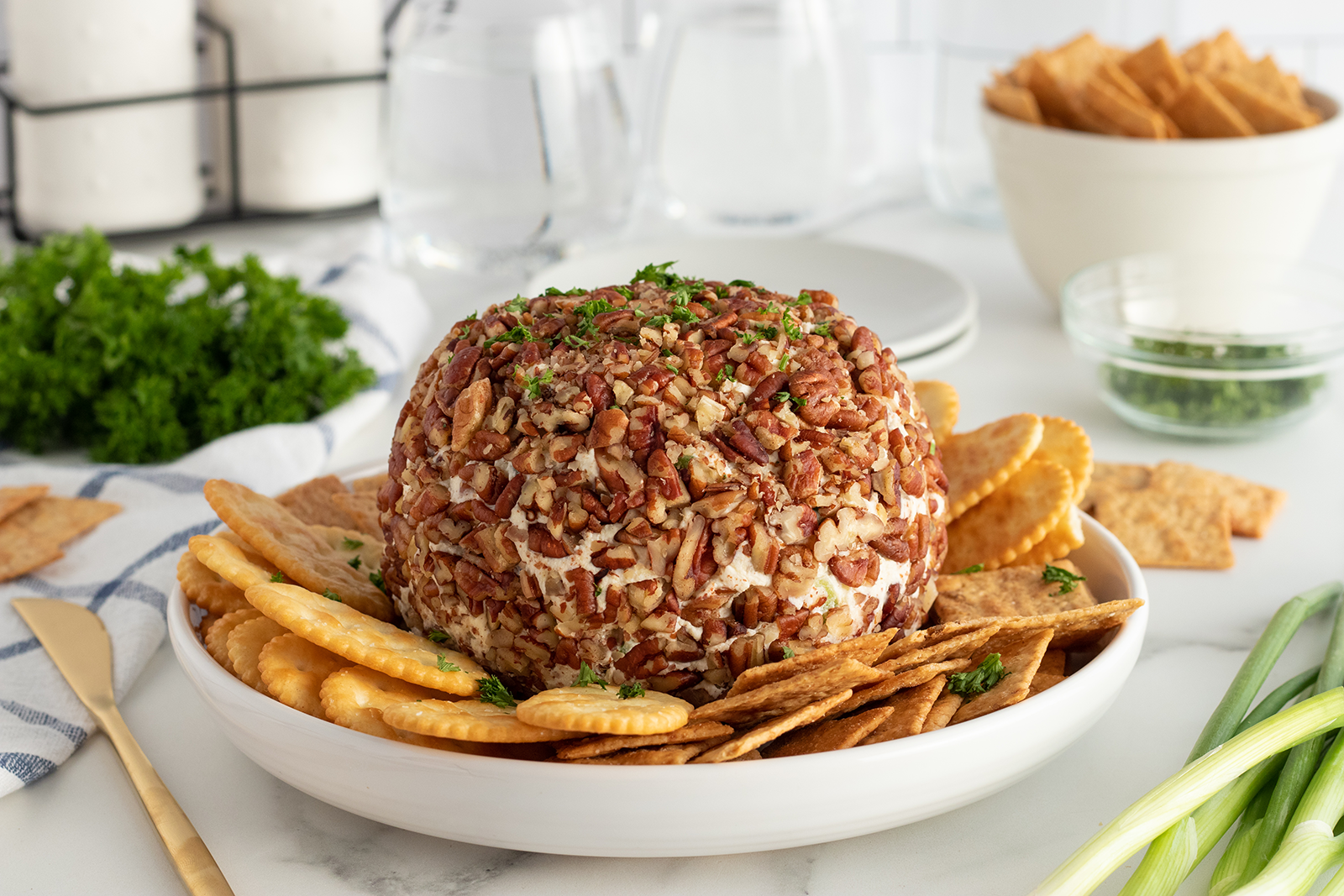A round cheese ball coated in chopped pecans sits on a white plate, surrounded by assorted crackers. Parsley garnish and green onions are nearby, with bowls of crackers and herbs in the background.